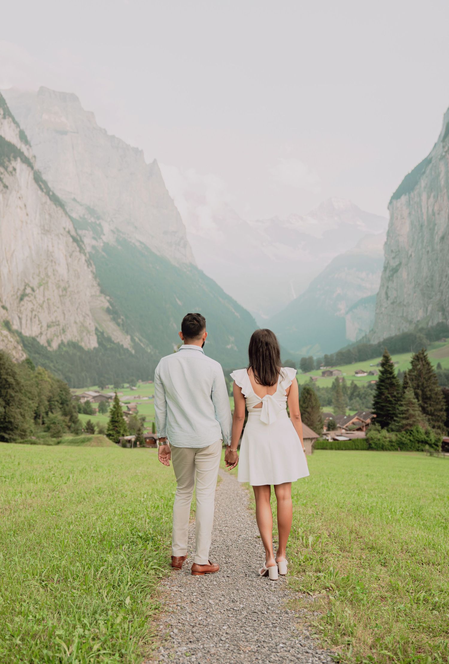 Romantic couple walking hand in hand toward misty Swiss Alps mountains on a gravel path.
