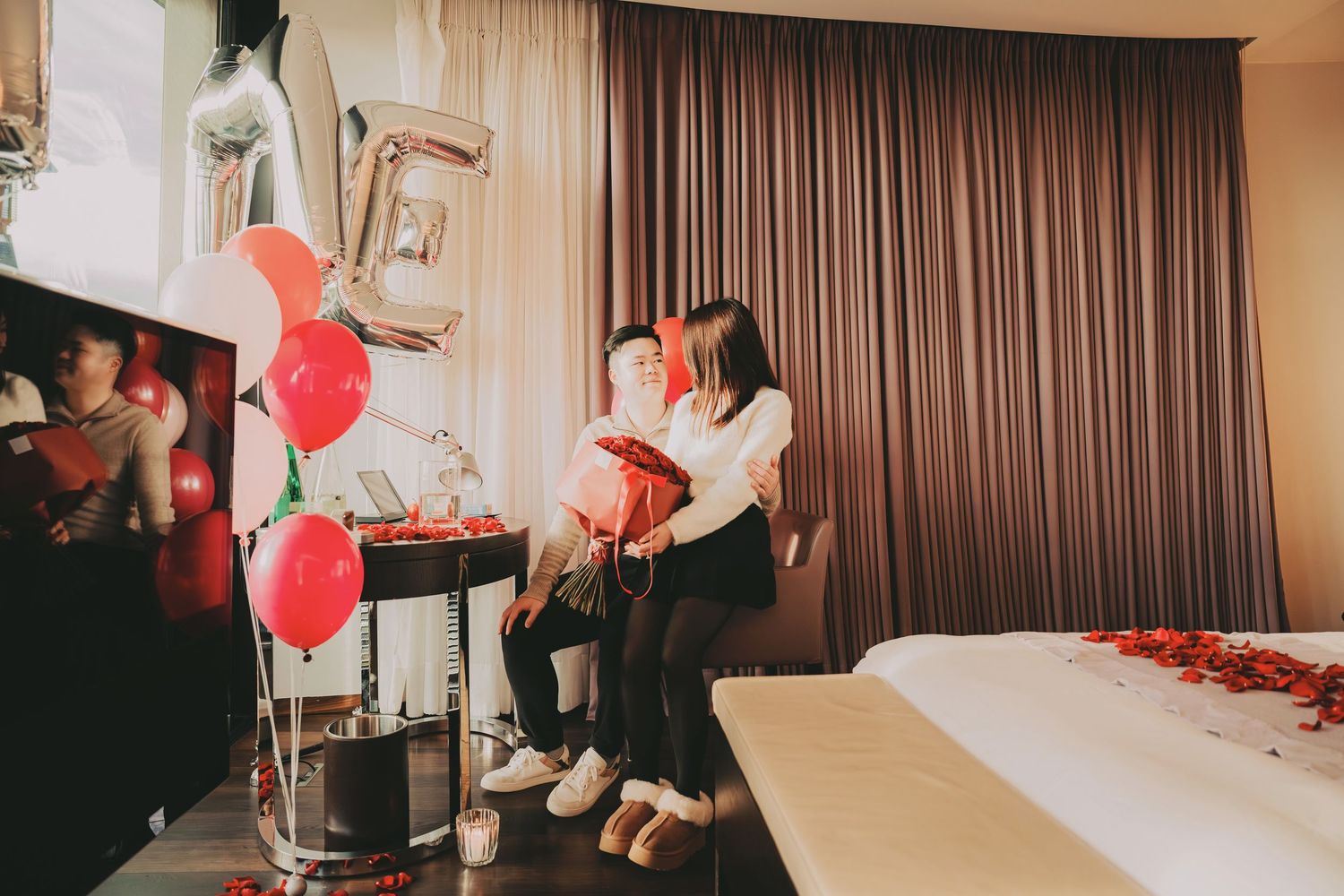 Asian couple sitting on a bed in a romantic hotel room decorated with red balloons and rose petals.