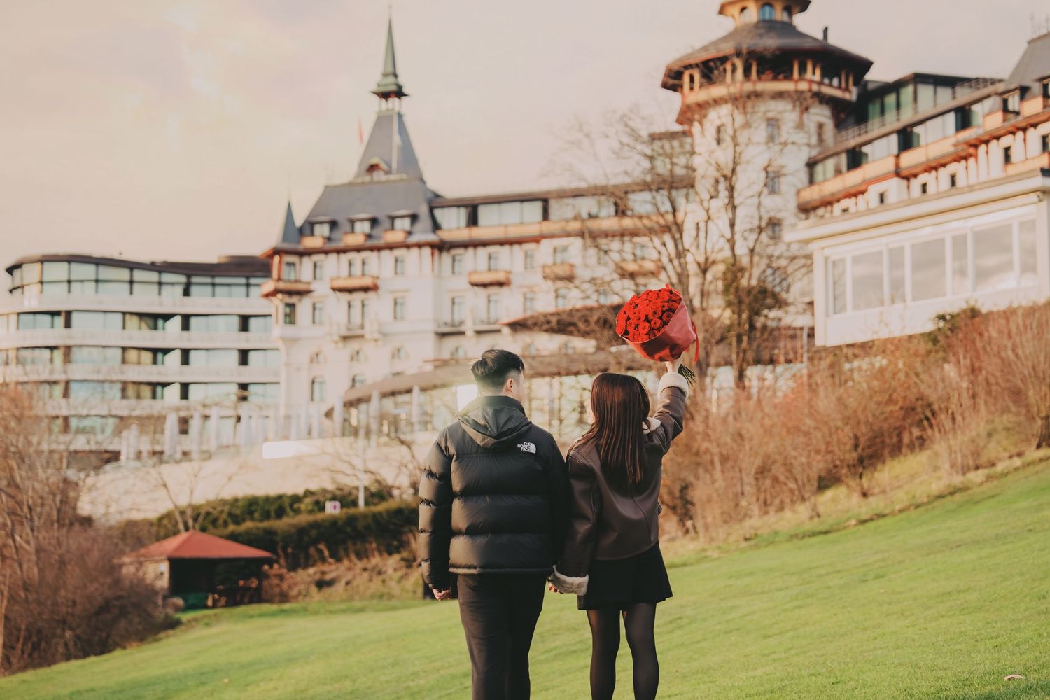 Romantic couple holding hands walking toward grand European hotel with towers on green lawn.