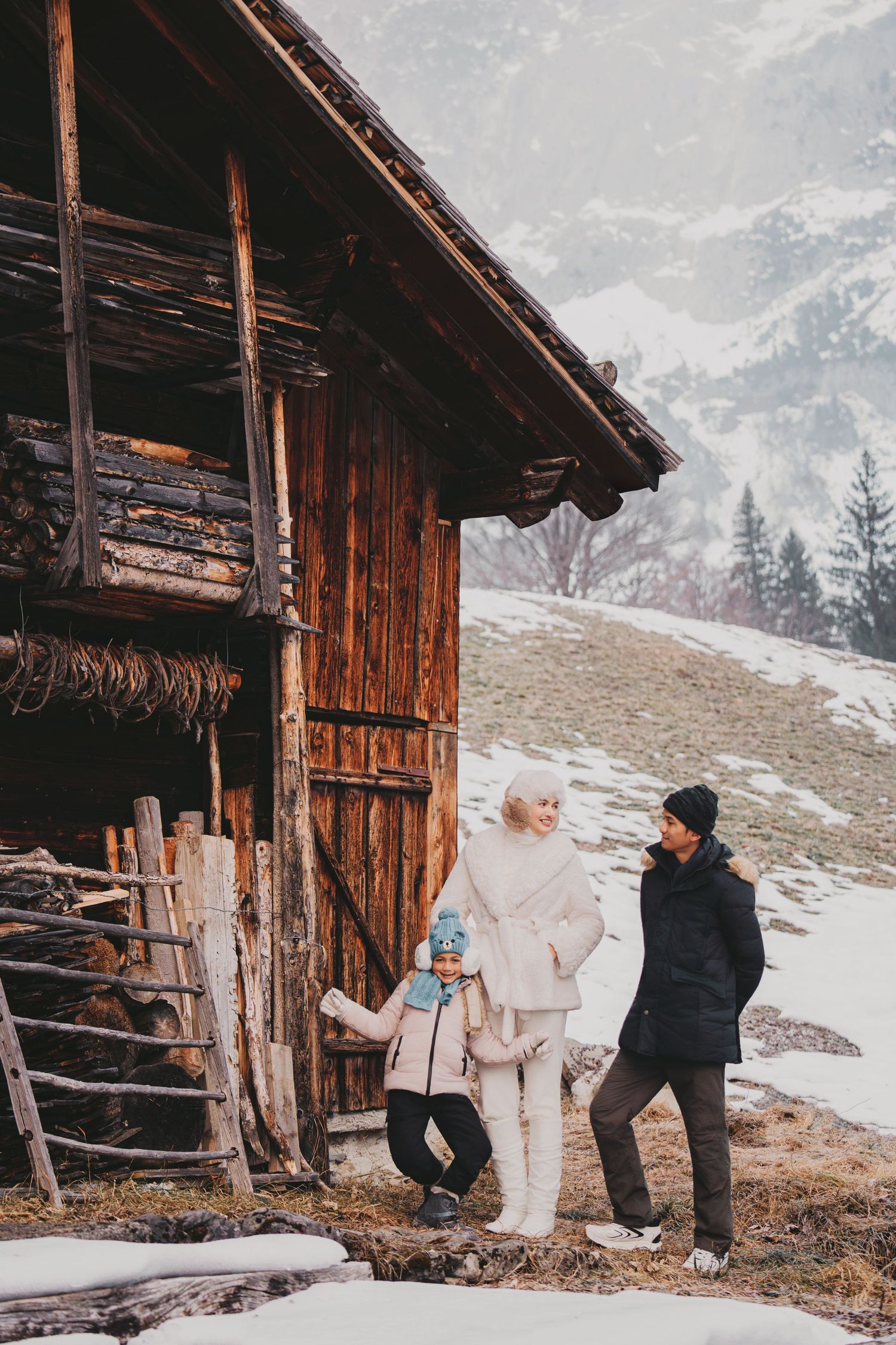 Family of three standing by a rustic wooden alpine barn on a snowy hillside.