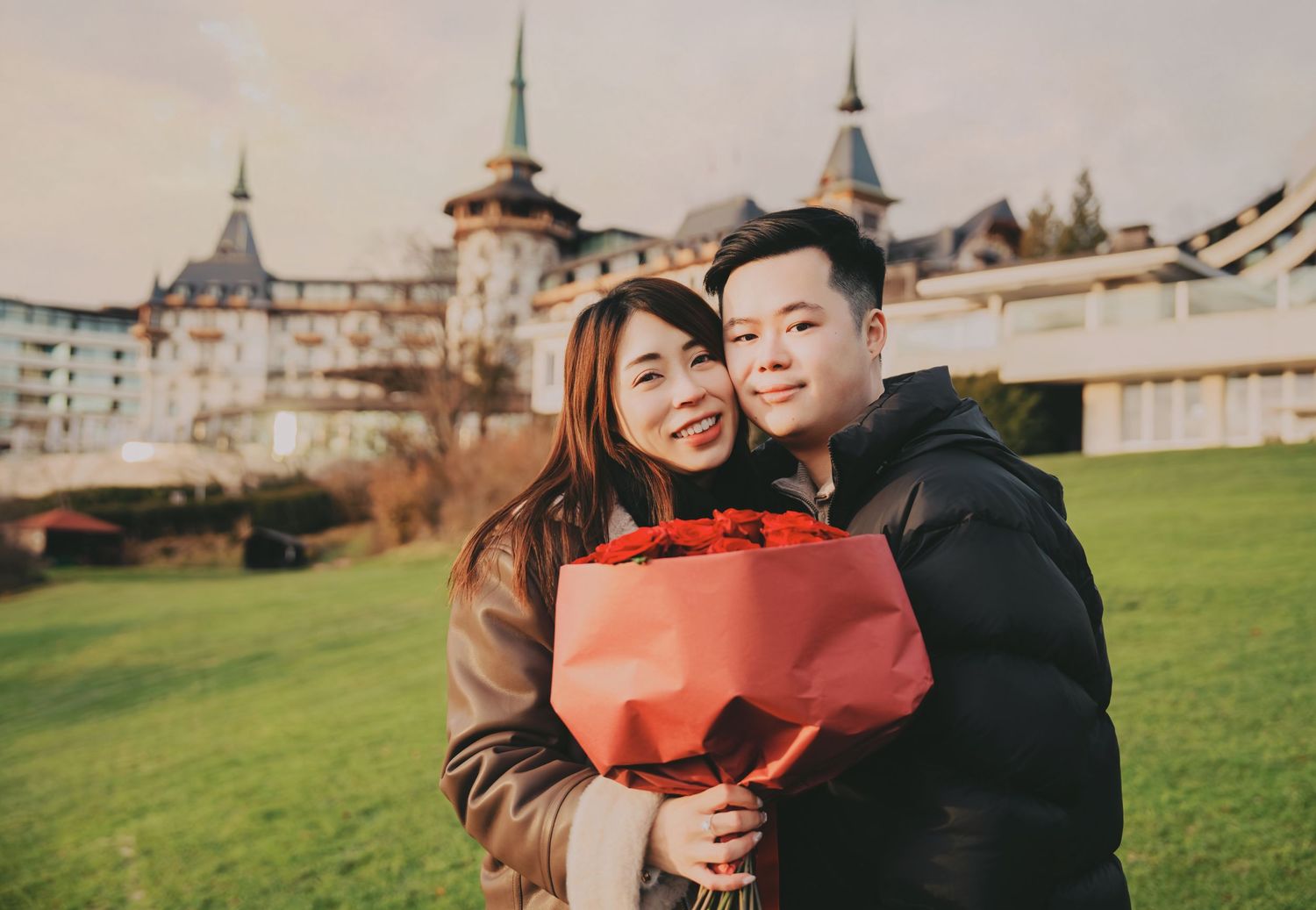 Happy Asian couple smiling holding red rose bouquet with castle hotel backdrop.