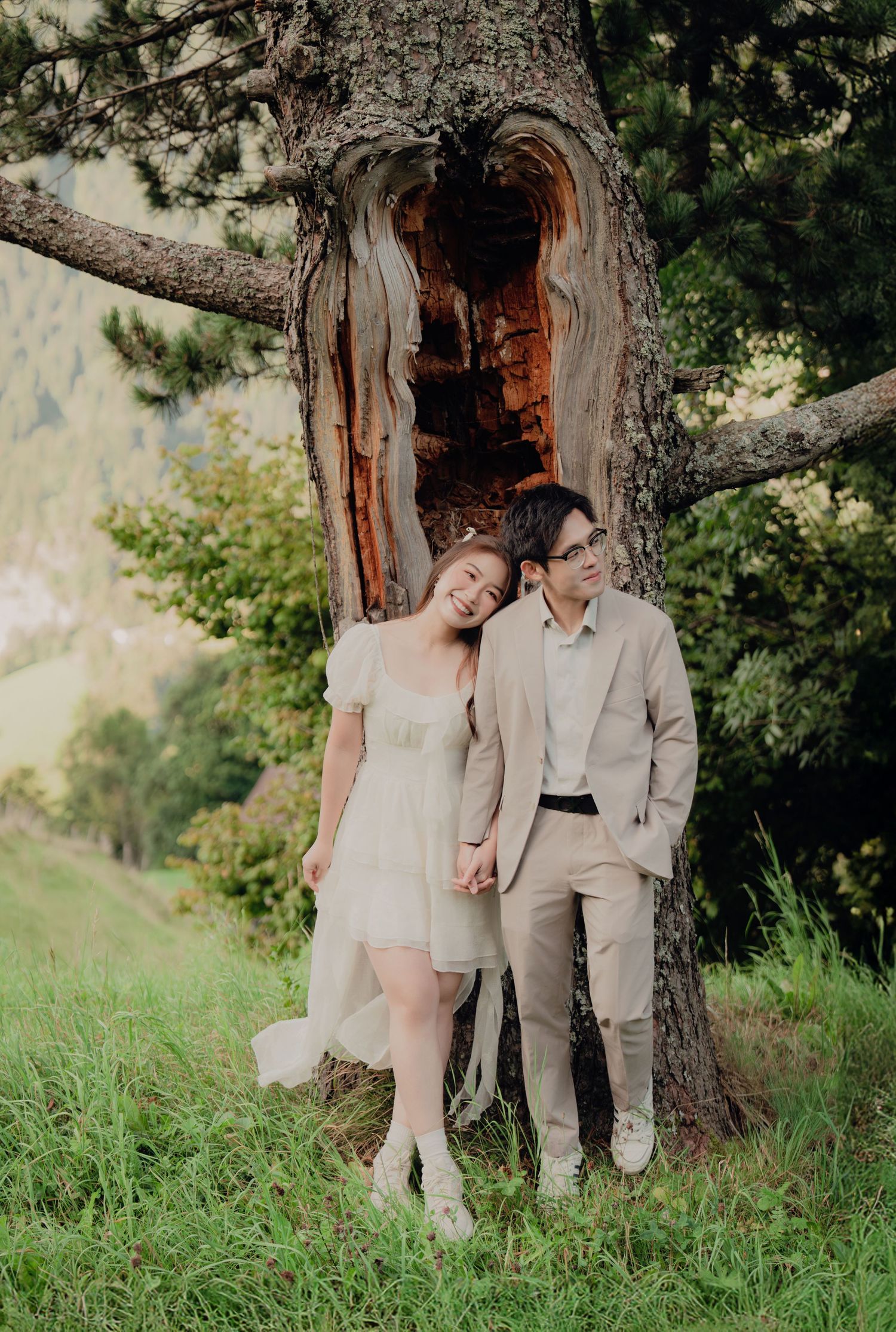 Happy couple leans against a gnarled pine tree on a grassy alpine slope with scenic mountain valley views.