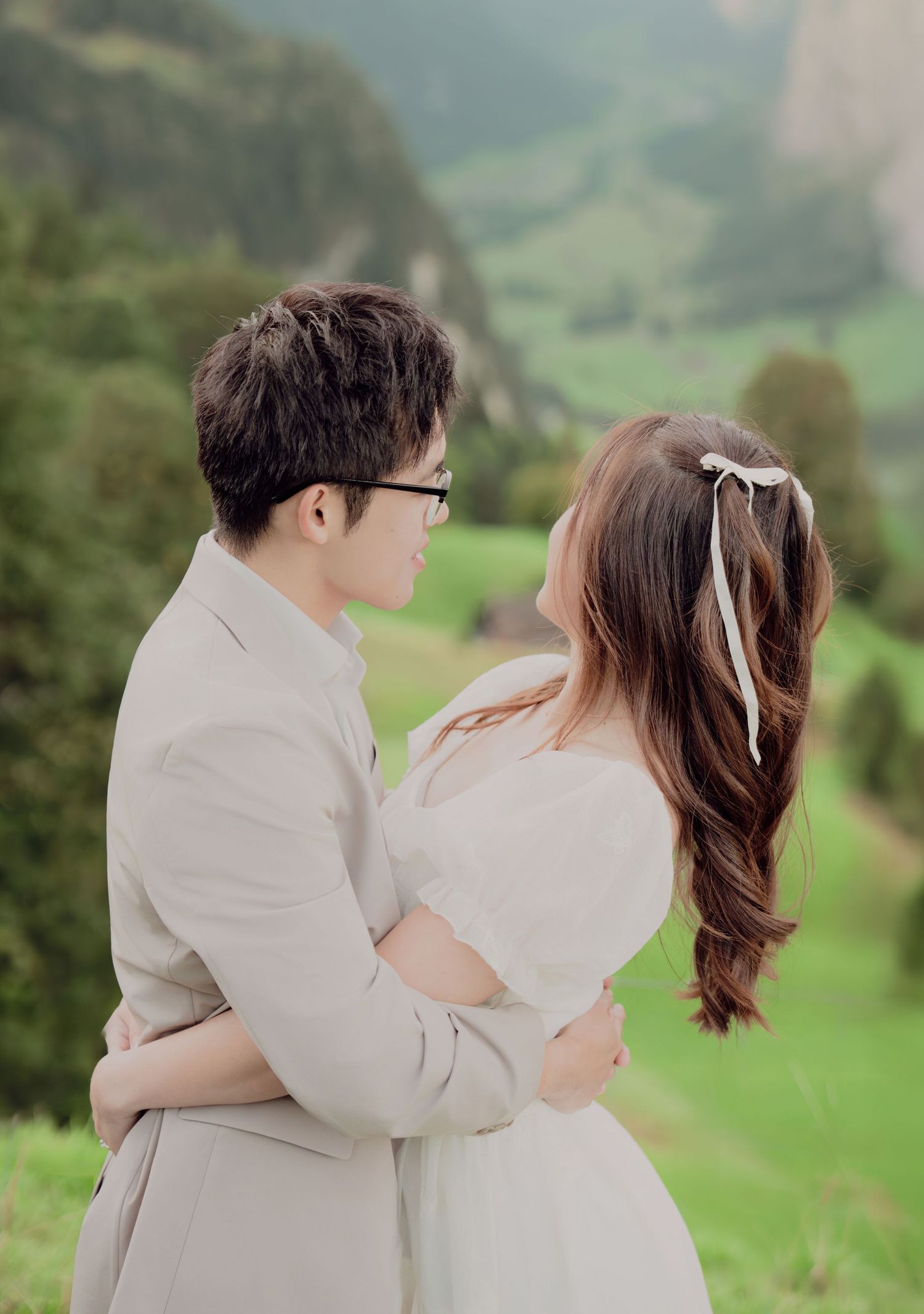 Couple in wedding attire holding each other and gazing into each other's eyes in a green alpine meadow.
