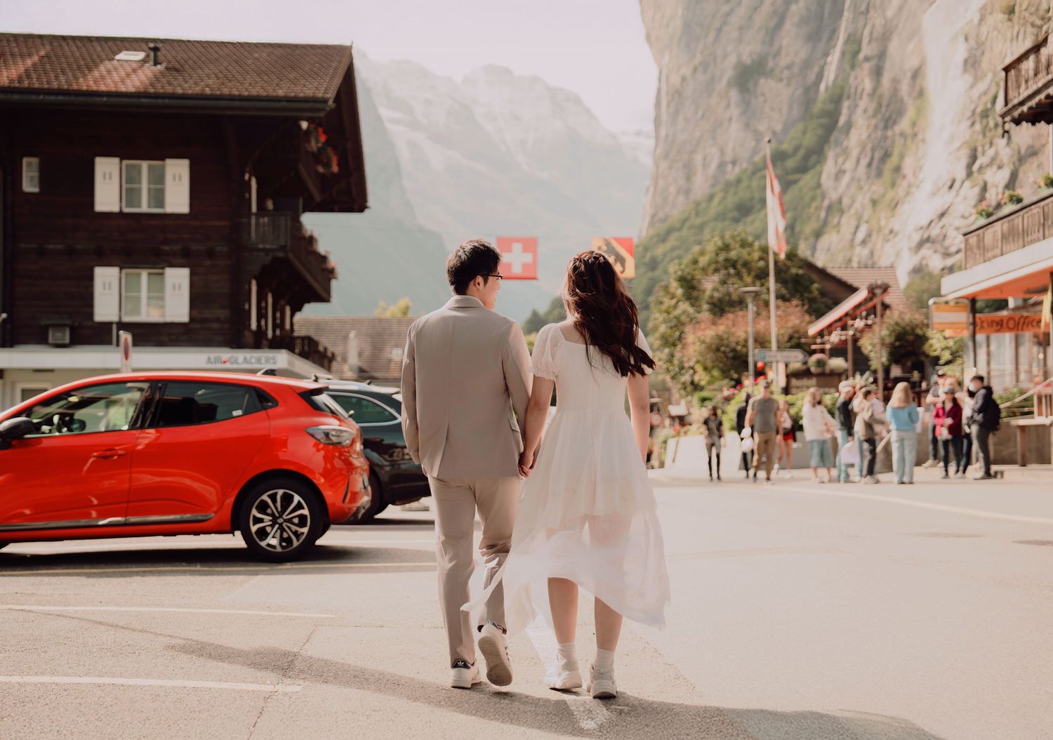 Couple walking hand in hand through Lauterbrunnen village street with red car and Swiss Alps backdrop.