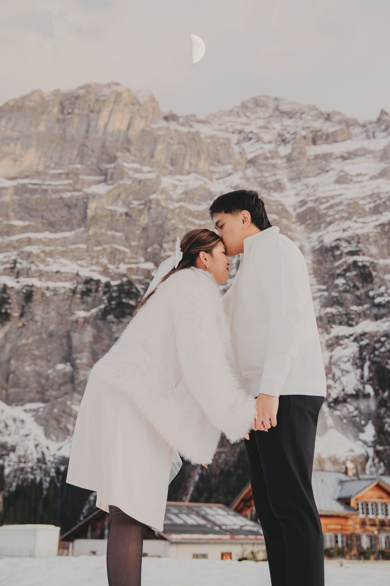 Couple sharing a tender kiss in snow with crescent moon and rocky mountain in background.