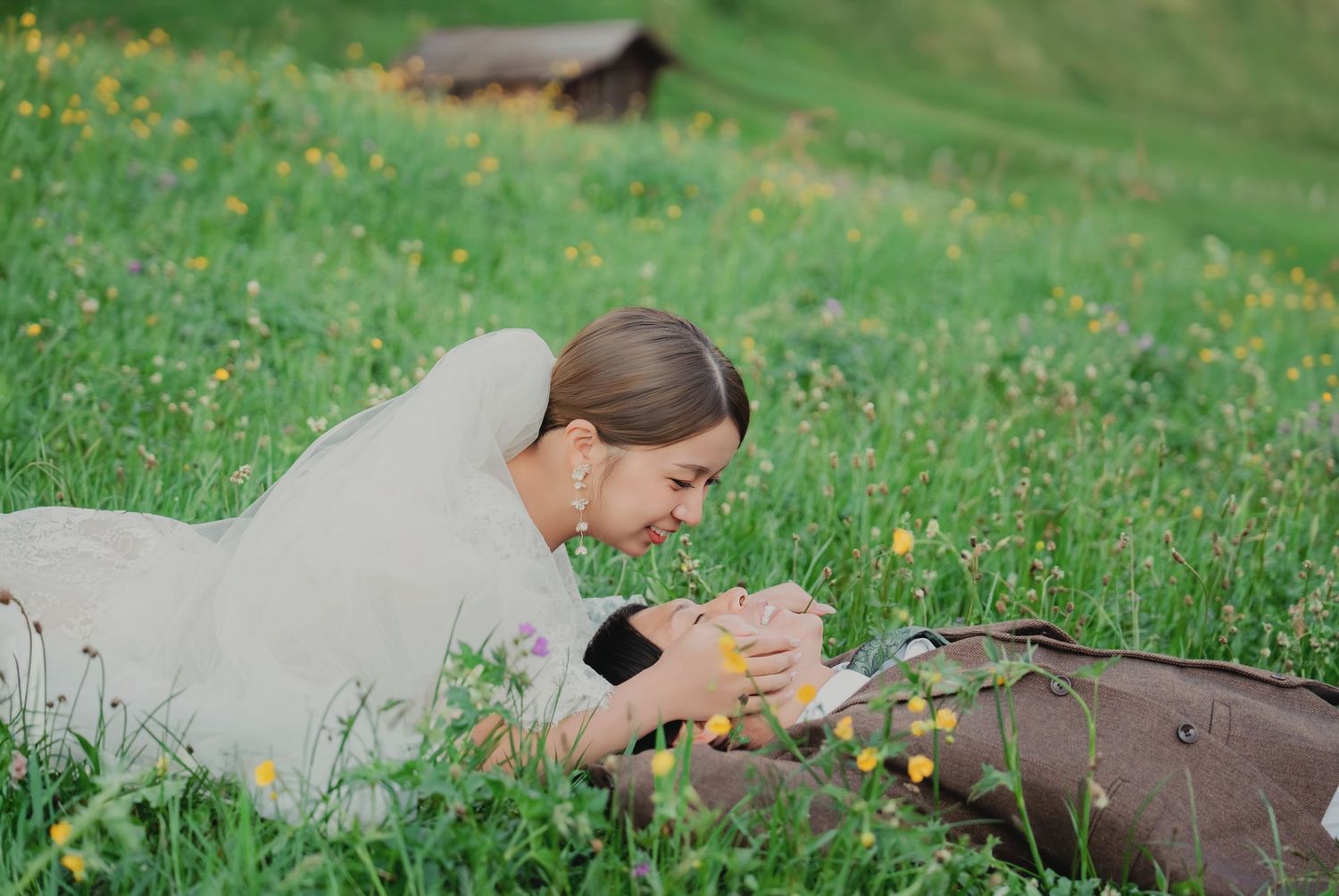 Bride lying in yellow wildflower meadow sharing tender moment with groom in Swiss Alps.