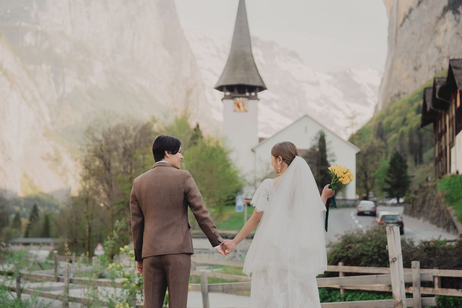 Bride and groom walking hand in hand toward a Swiss alpine church with misty mountain scenery.