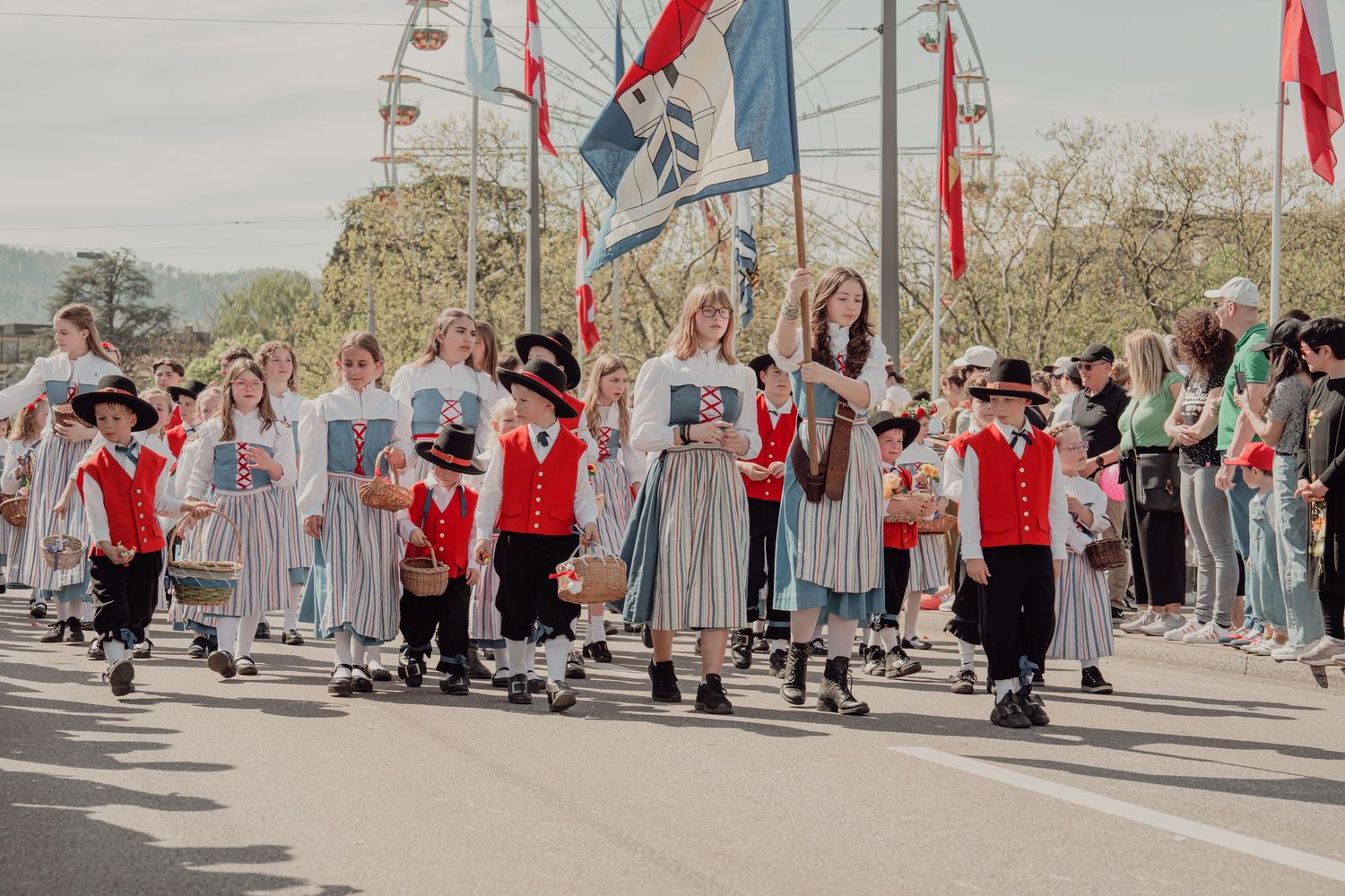 Children and adults in traditional Swiss folk costumes march in a colorful cultural parade with flags.