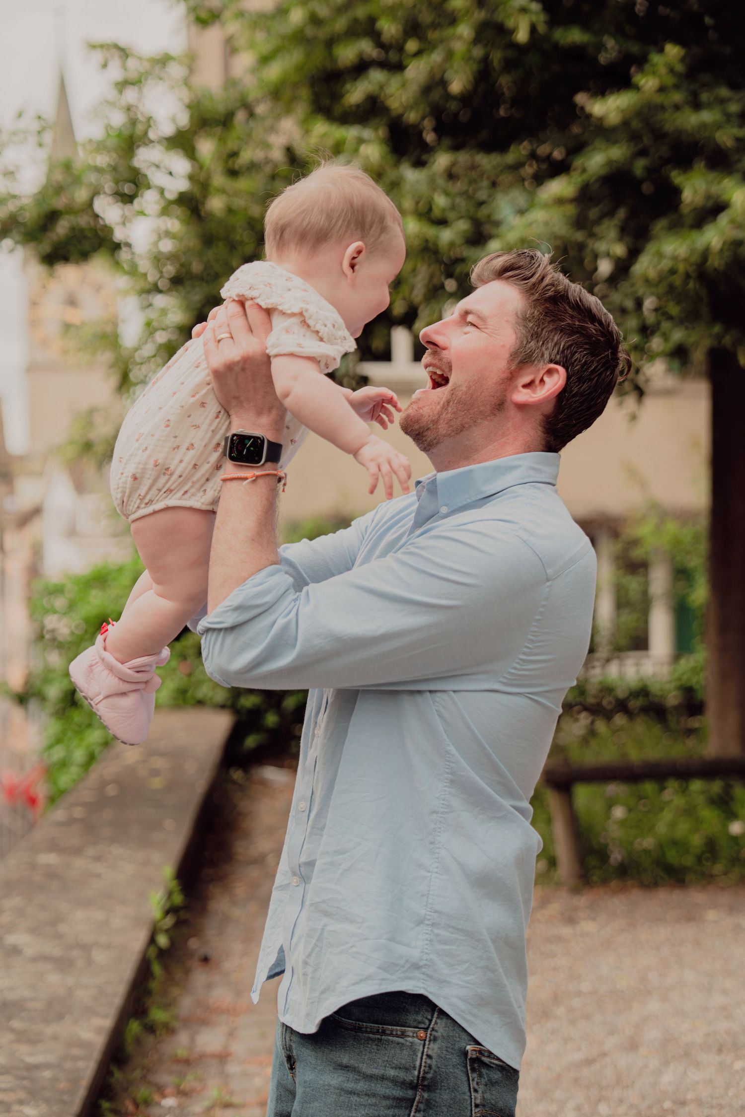 Father joyfully lifting smiling baby in floral outfit outdoors in a lush green European setting.