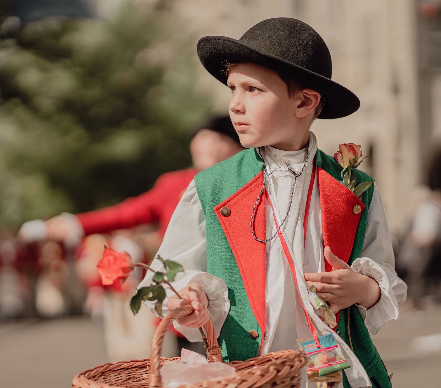 Young boy in traditional costume holds a rose and basket during a colorful cultural street parade.