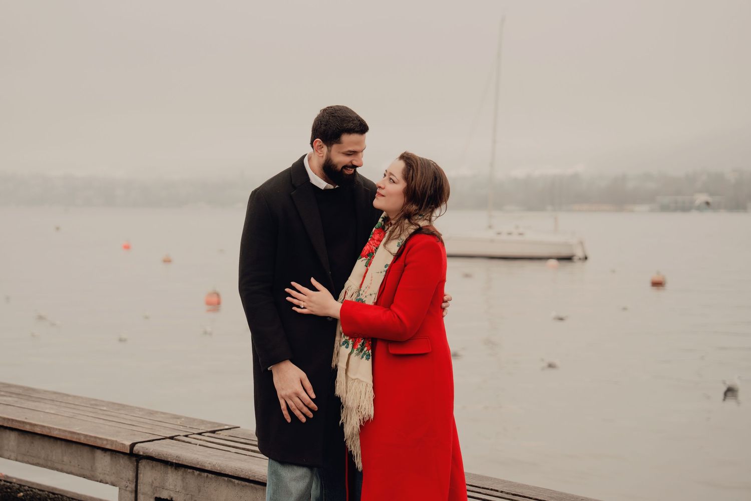 Couple gazing at each other romantically by a misty lake, woman in red coat and patterned scarf.
