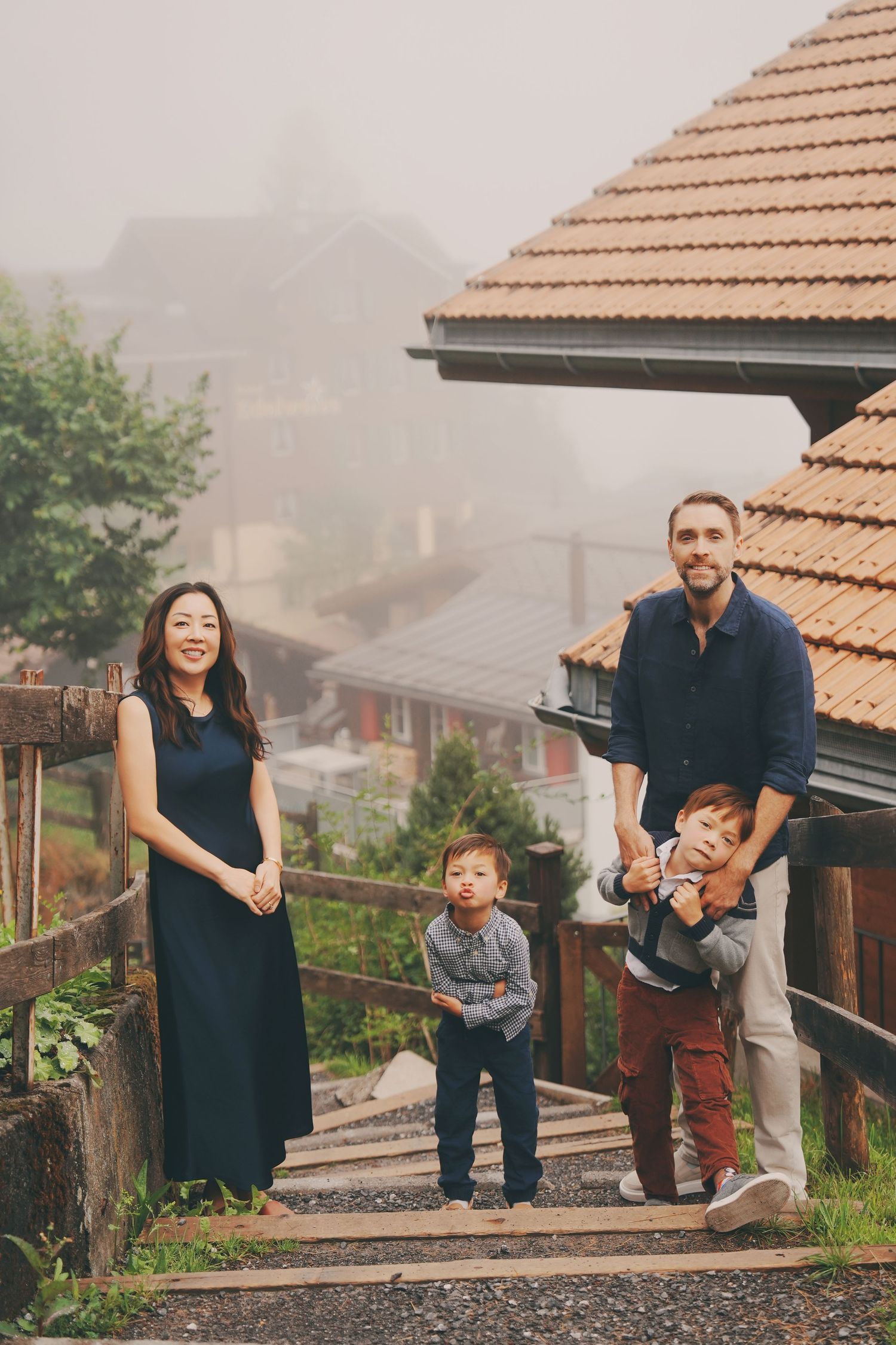 A family of four enjoys a misty Swiss alpine village setting with a father playfully holding his son.