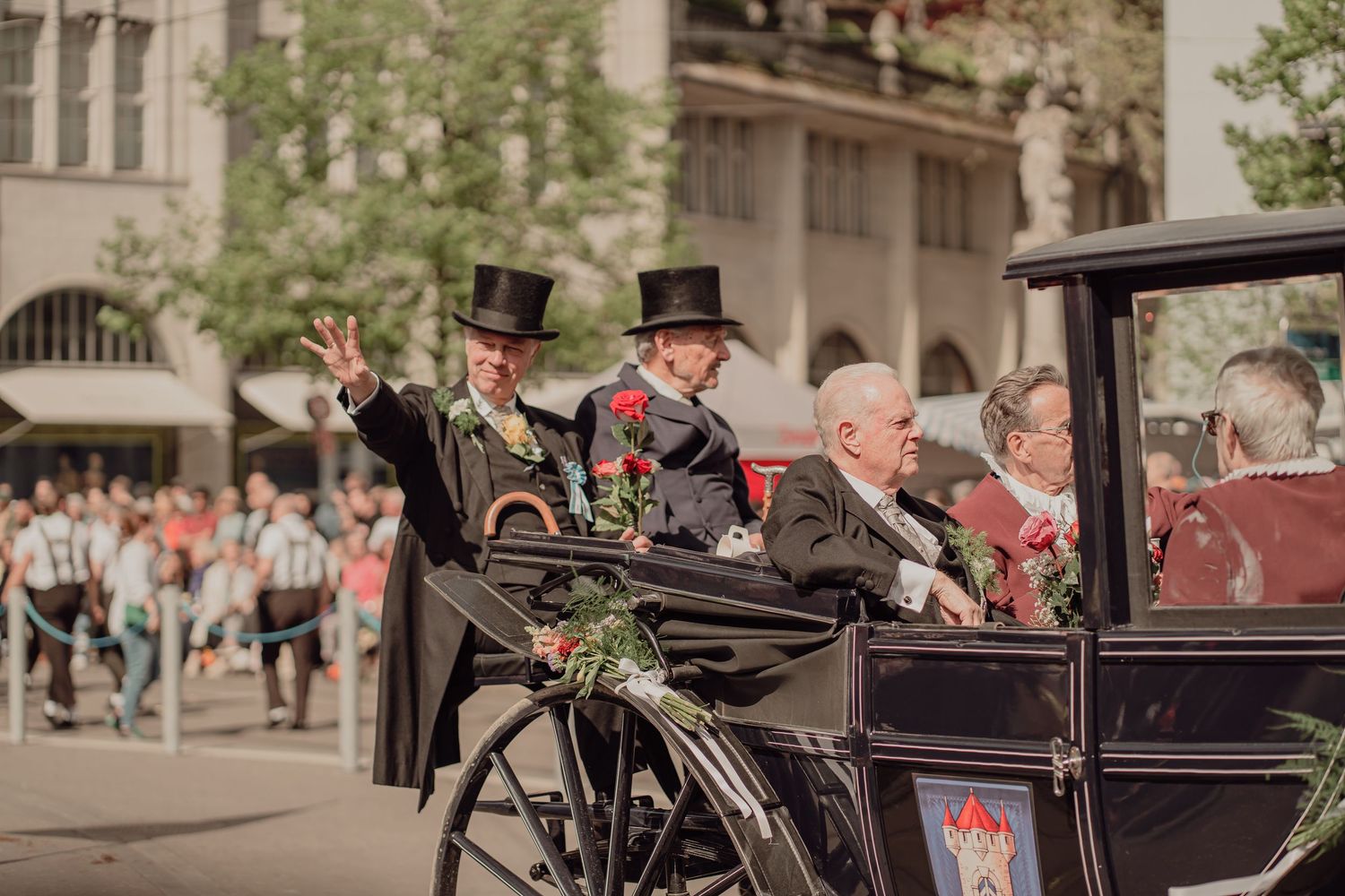 Two men in top hats wave from a black carriage during a festive outdoor parade.