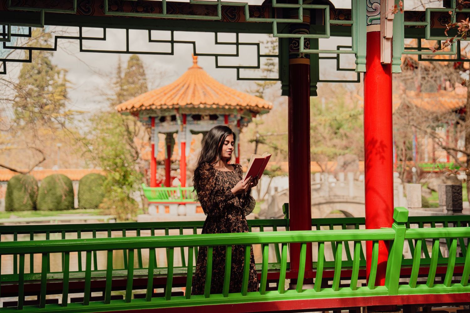 Woman in floral dress reads book on ornate green bridge in traditional Chinese garden pavilion setting.