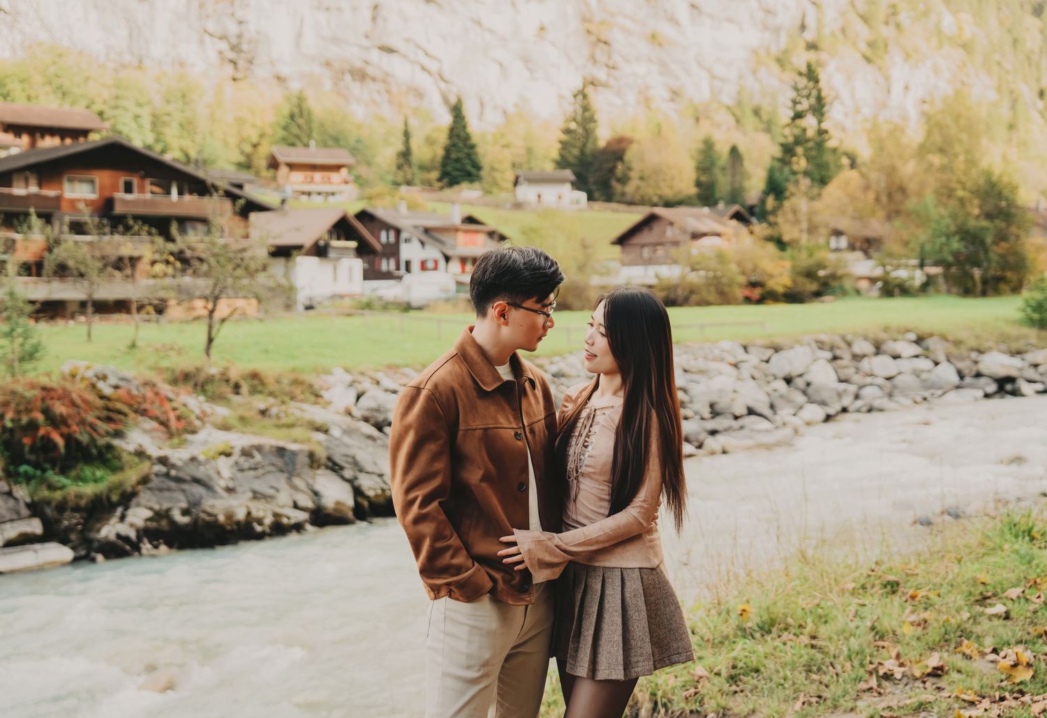 Asian couple sharing a romantic moment beside a turquoise alpine river in a scenic Swiss mountain village.