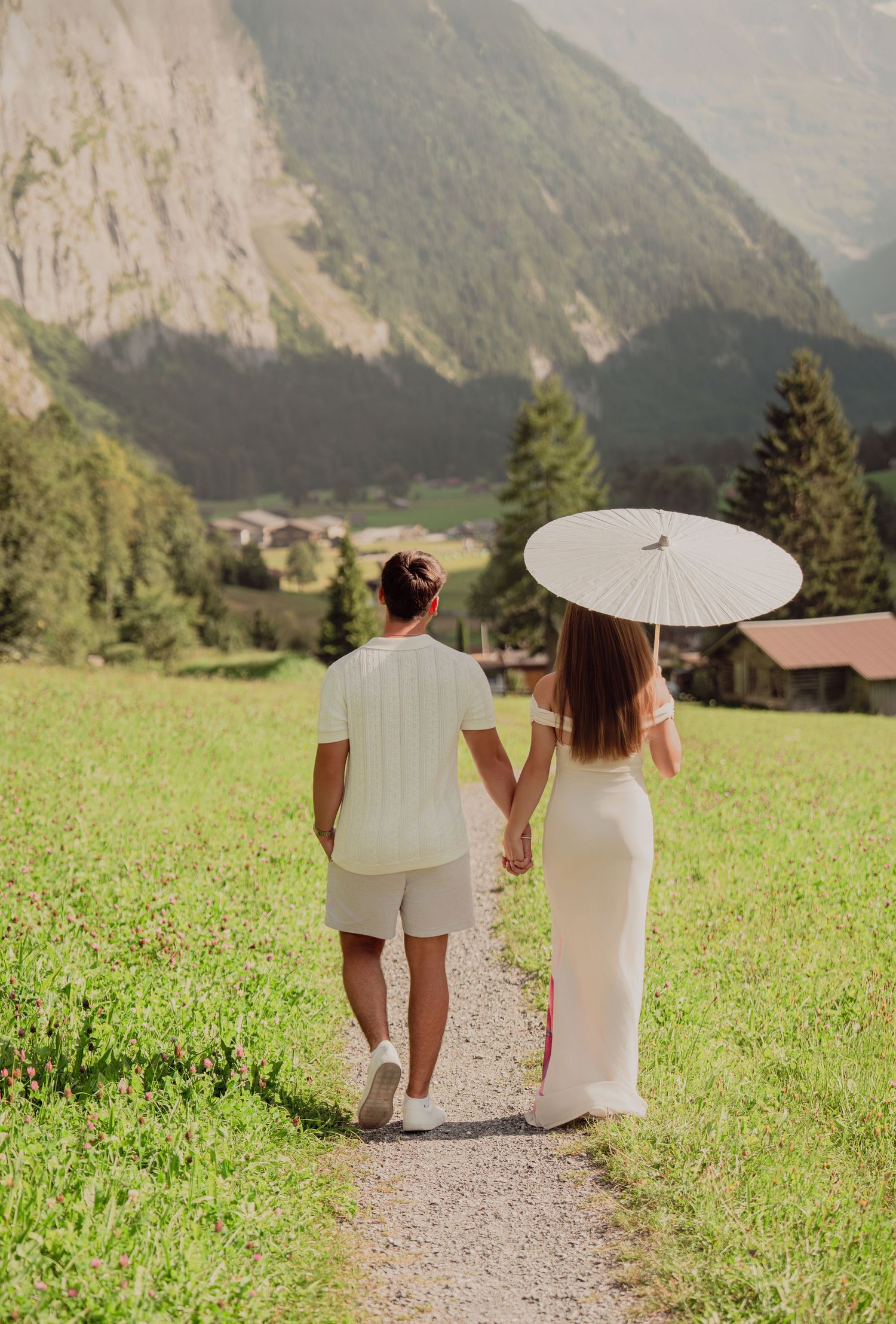 Couple walking hand in hand on a meadow path in a Swiss alpine valley, woman holding a white parasol.
