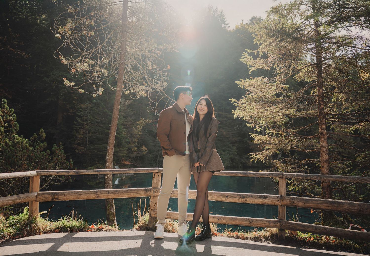 Couple standing together on a sunlit wooden bridge with a blue lake and lush green forest behind them.