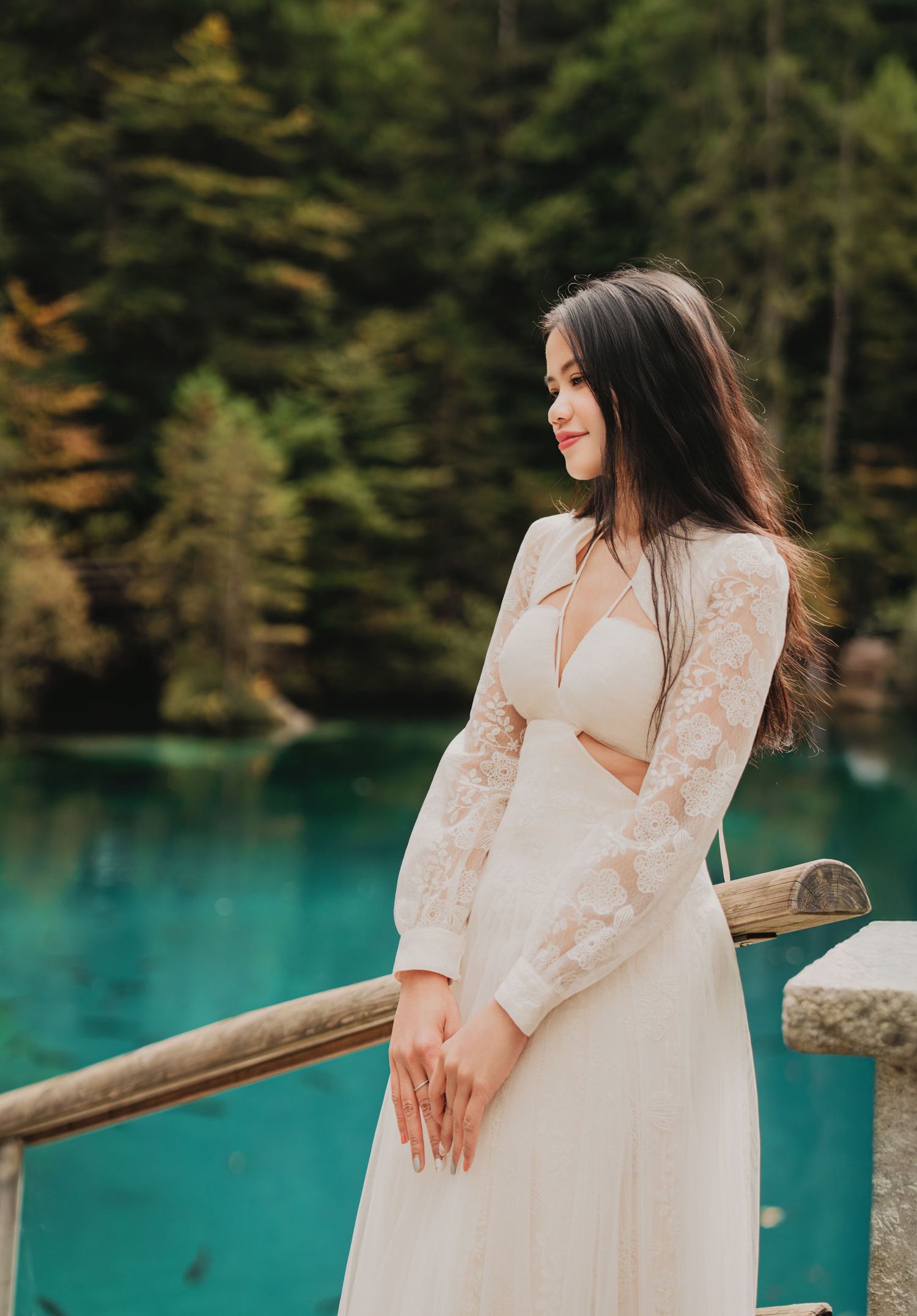 Young woman in white lace dress gazes down by turquoise lake surrounded by forest.