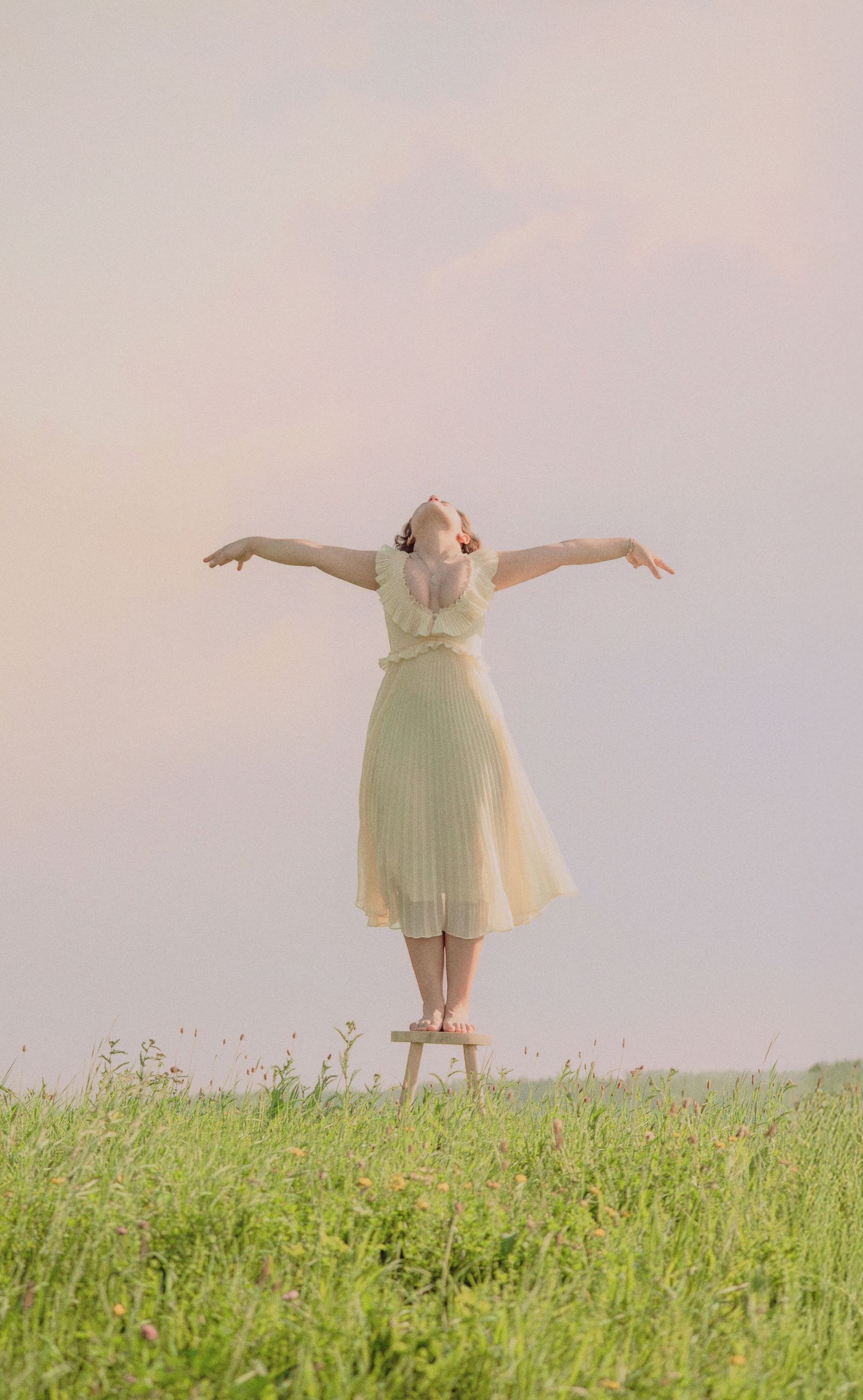 Woman in pale yellow dress standing on stool with arms outstretched in green meadow at dusk.