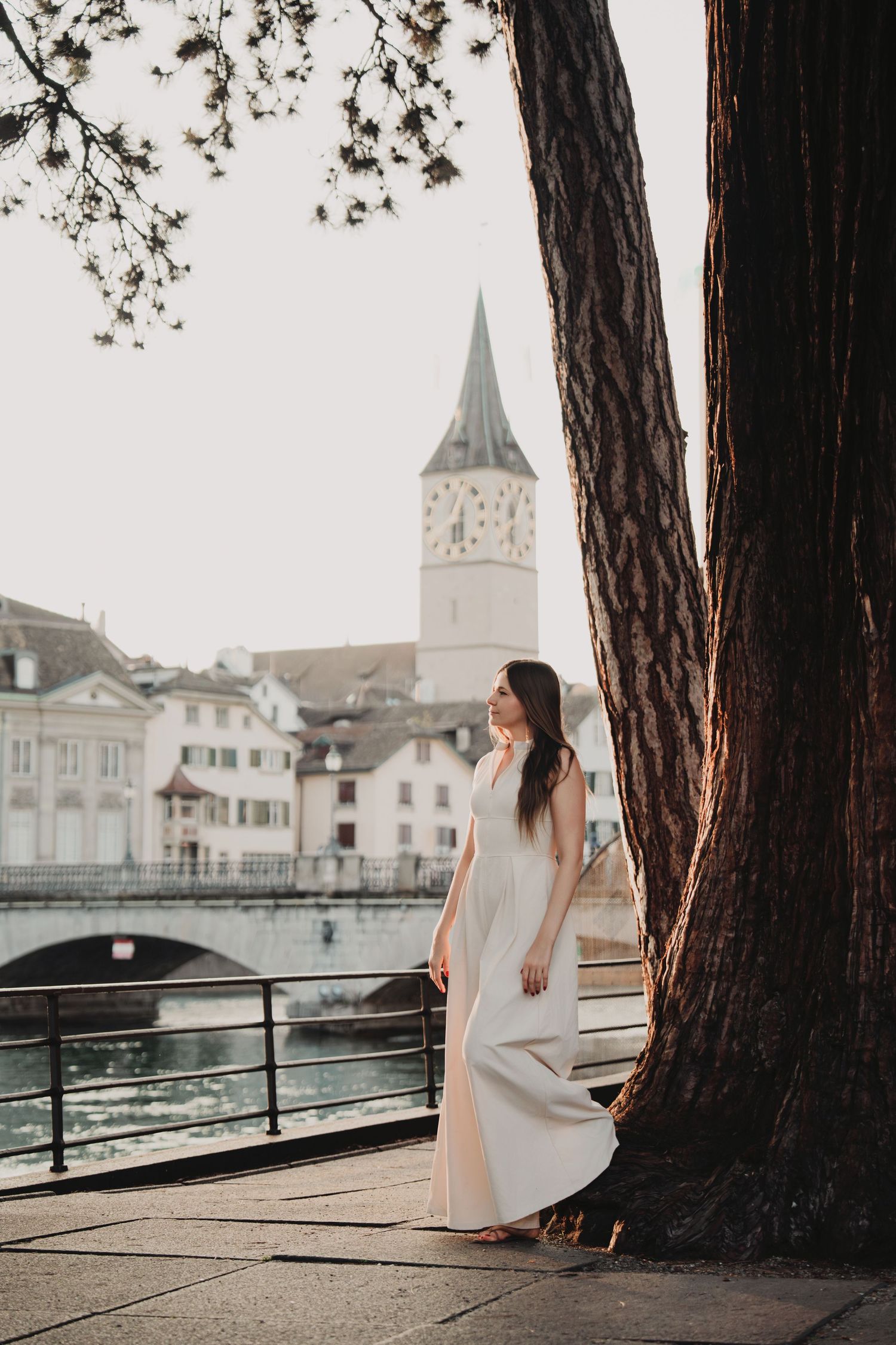 Woman in white maxi dress posing by large tree along Zurich river with St. Peter church tower view.