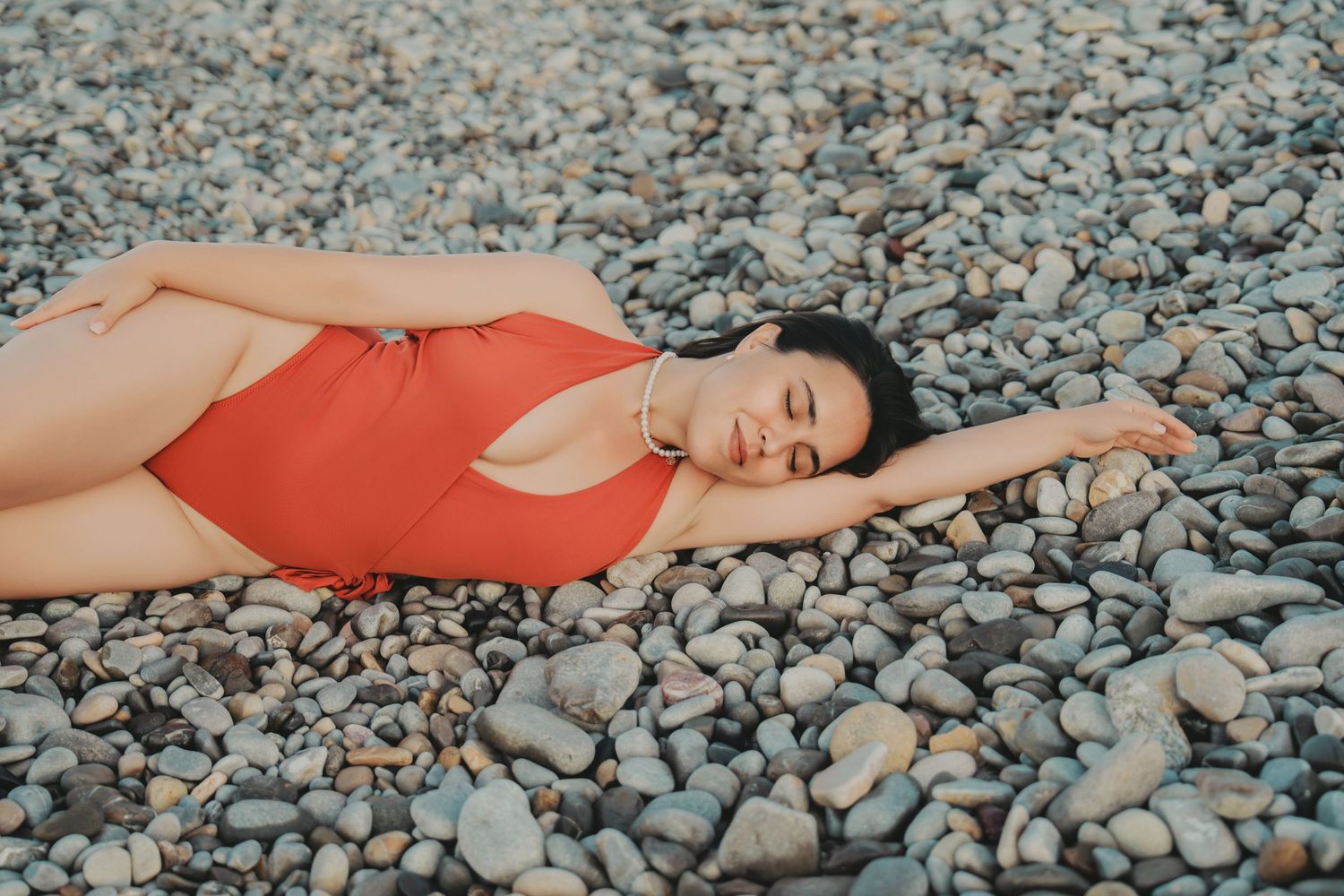 Woman in red swimsuit lying on pebble beach with eyes closed relaxing.
