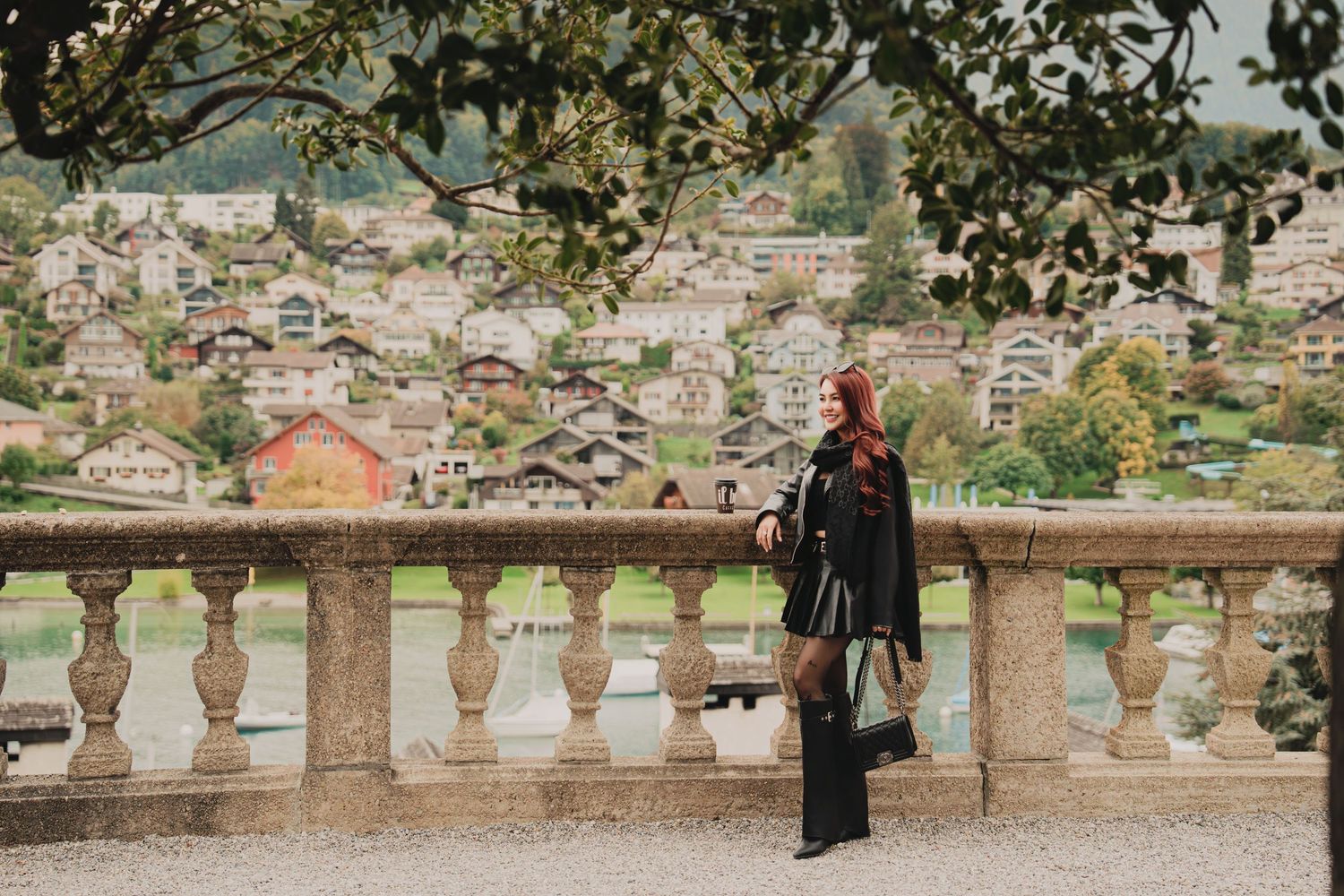 Red-haired woman in black outfit and tall boots stands at ornate stone balustrade with Swiss mountain village backdrop.