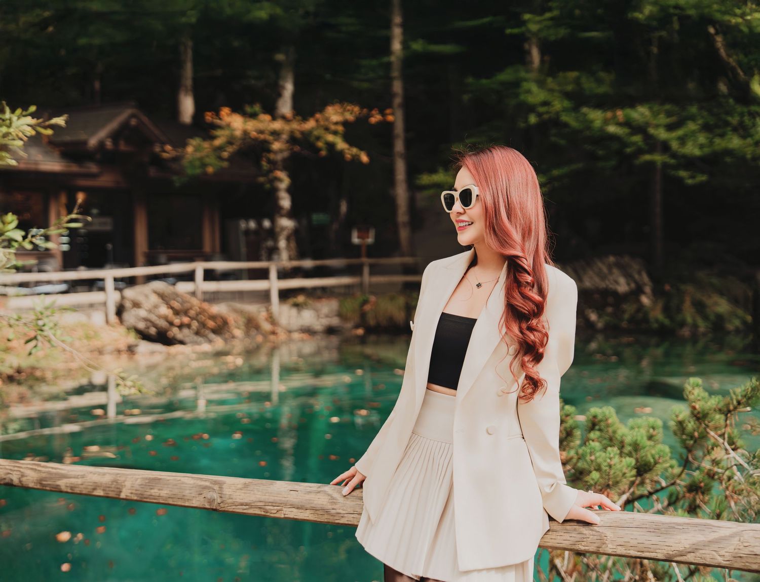 Woman in white blazer and sunglasses smiling by turquoise lake in autumn forest.