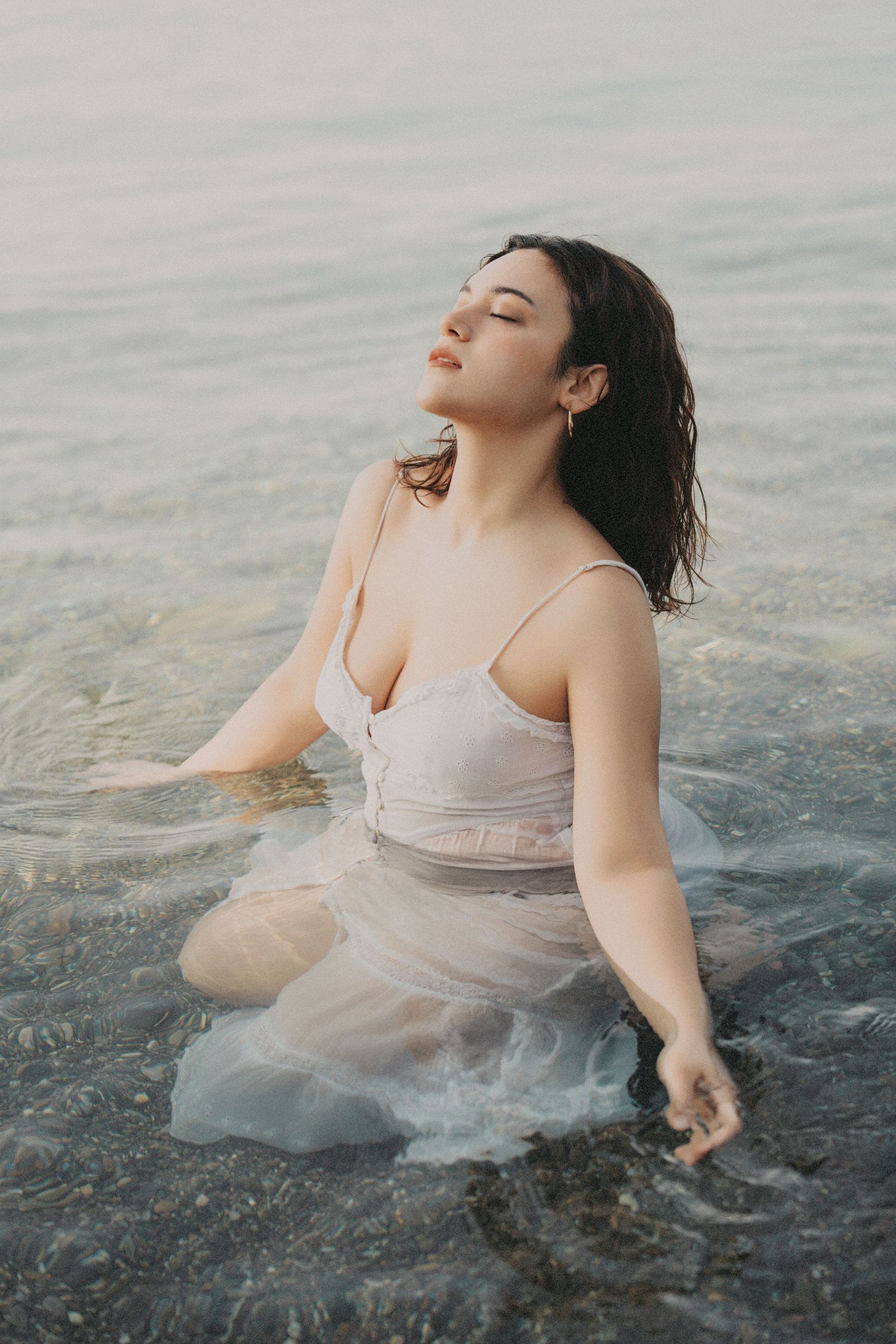 Woman in wet white dress kneeling in shallow water eyes closed.