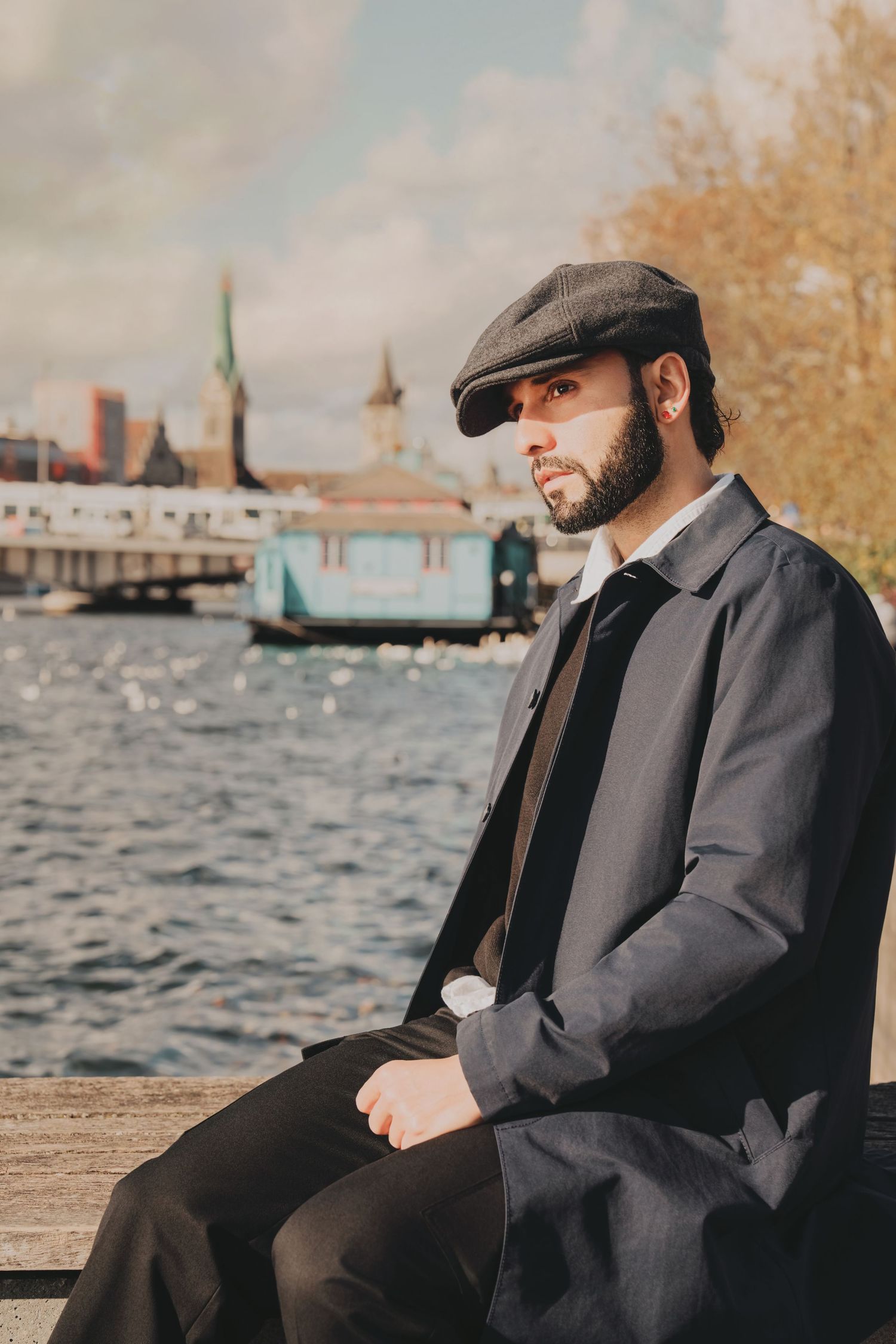 Bearded man in dark trench coat and flat cap sits pensively by a river with city skyline behind.
