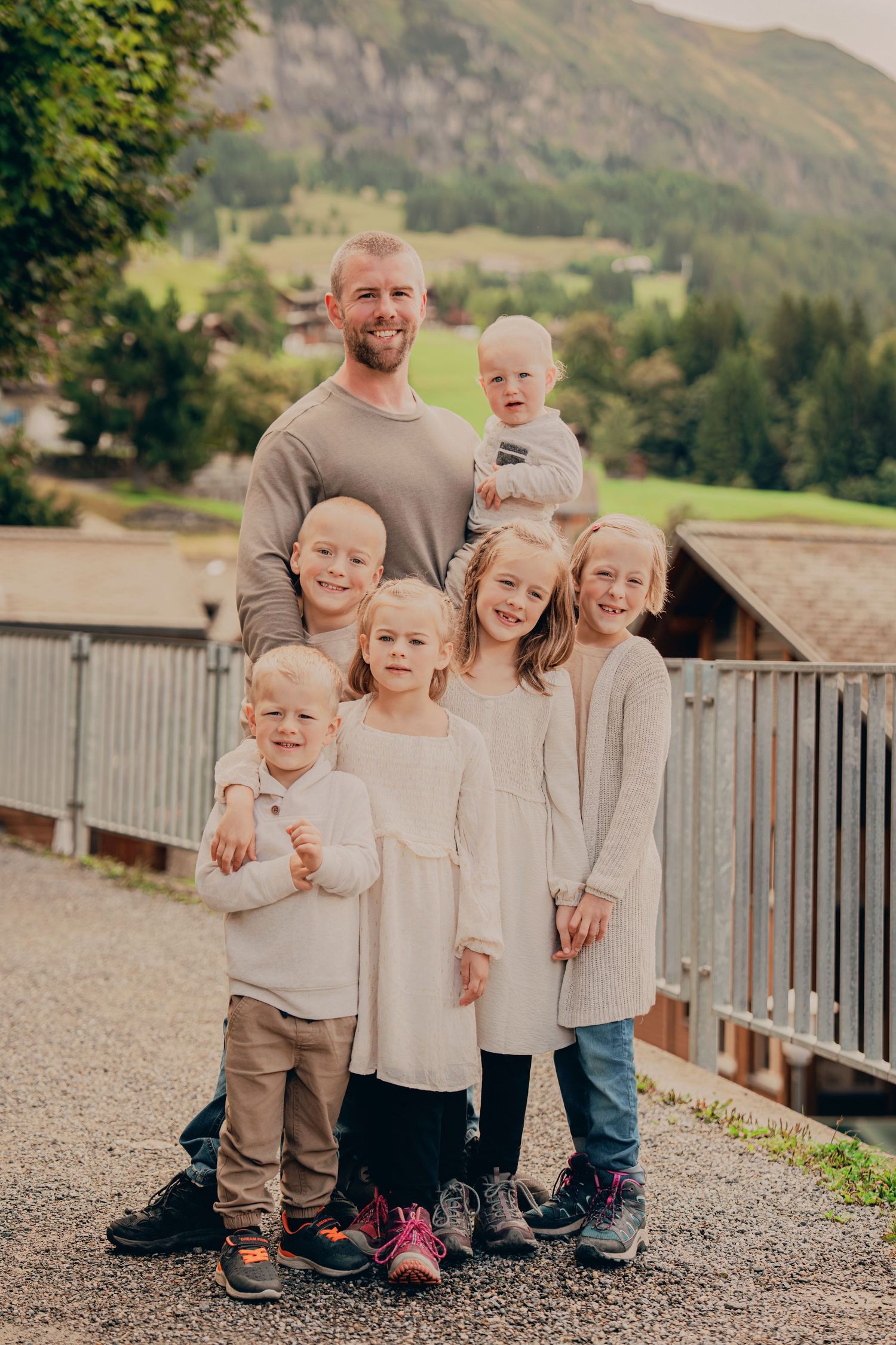 A father smiles proudly with his six young children on a bridge in an alpine village.