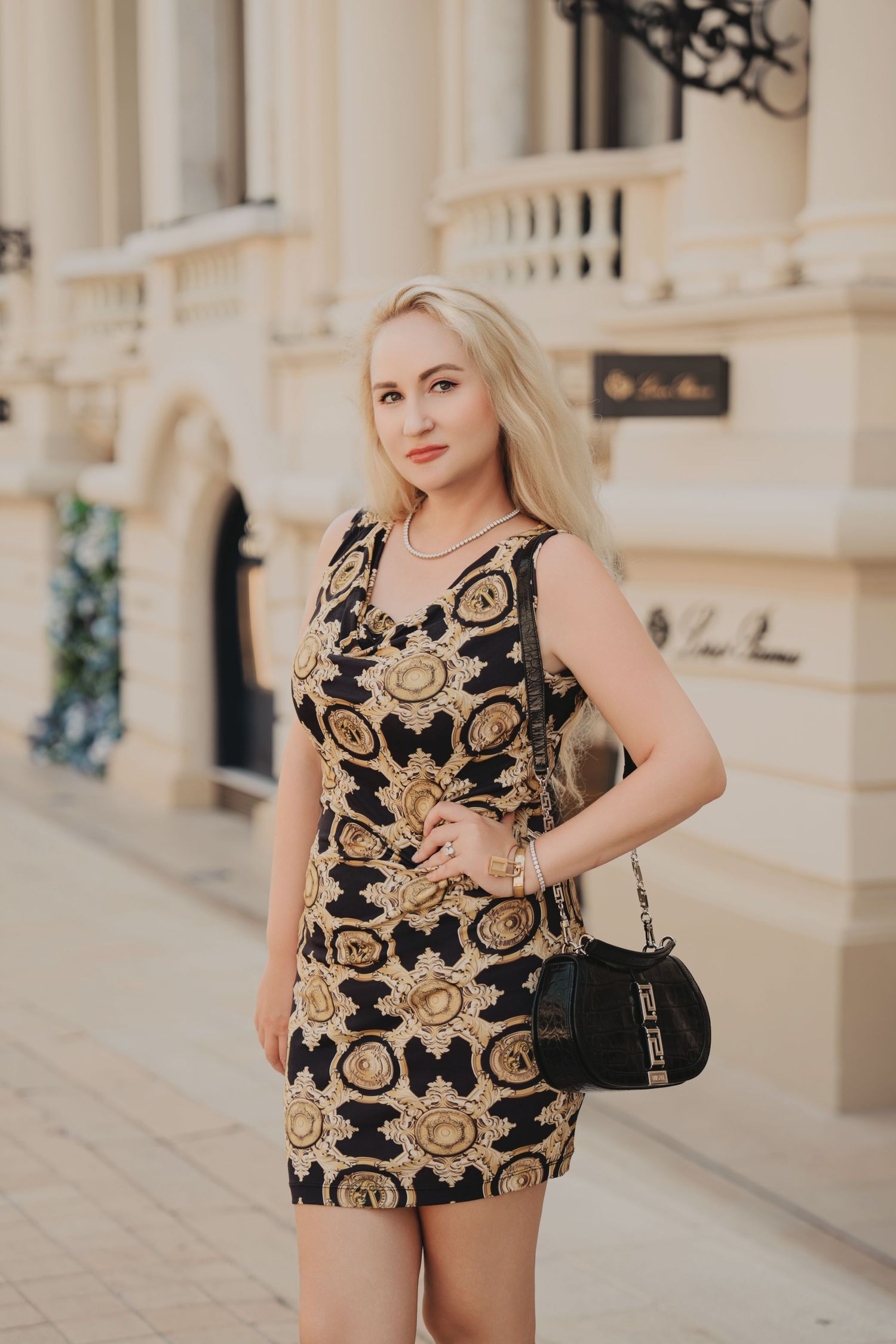 Blonde woman in black and gold Versace-style mini dress with handbag posing on an elegant European street.
