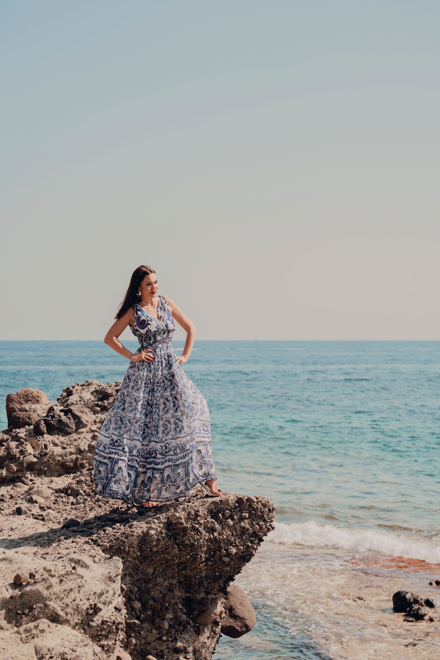 Woman in blue and white patterned maxi dress standing on coastal rocks by the sea.