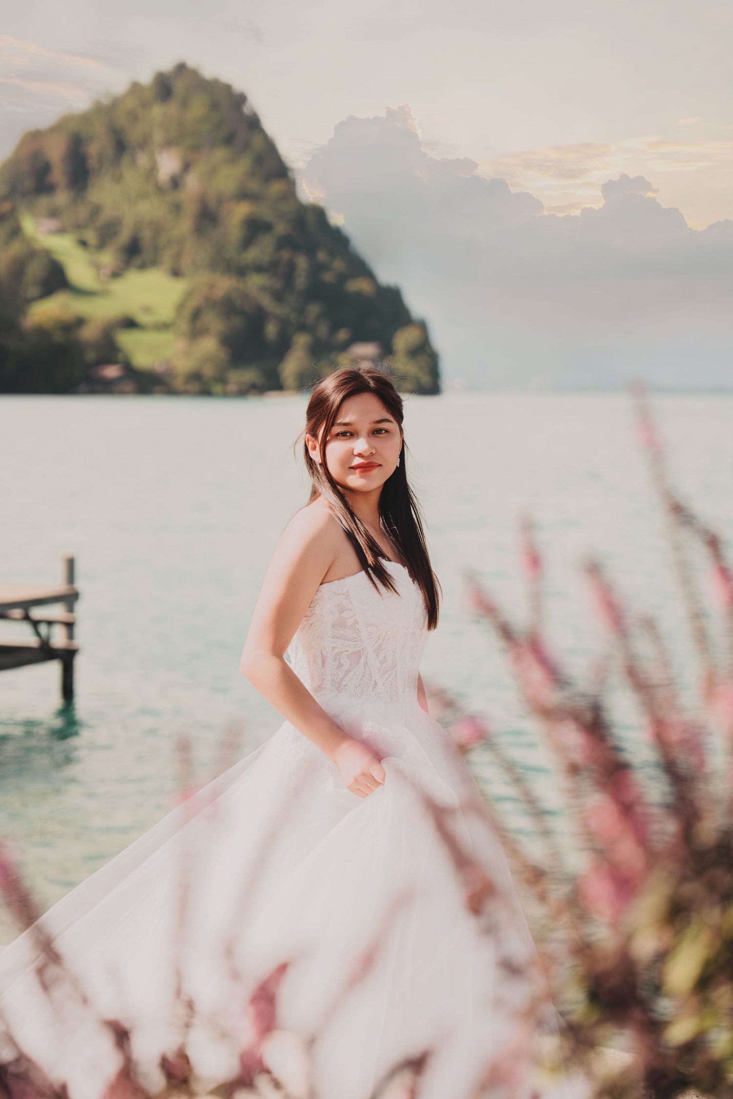 Bride in white strapless gown poses by turquoise alpine lake with mountain backdrop and pink wildflowers.