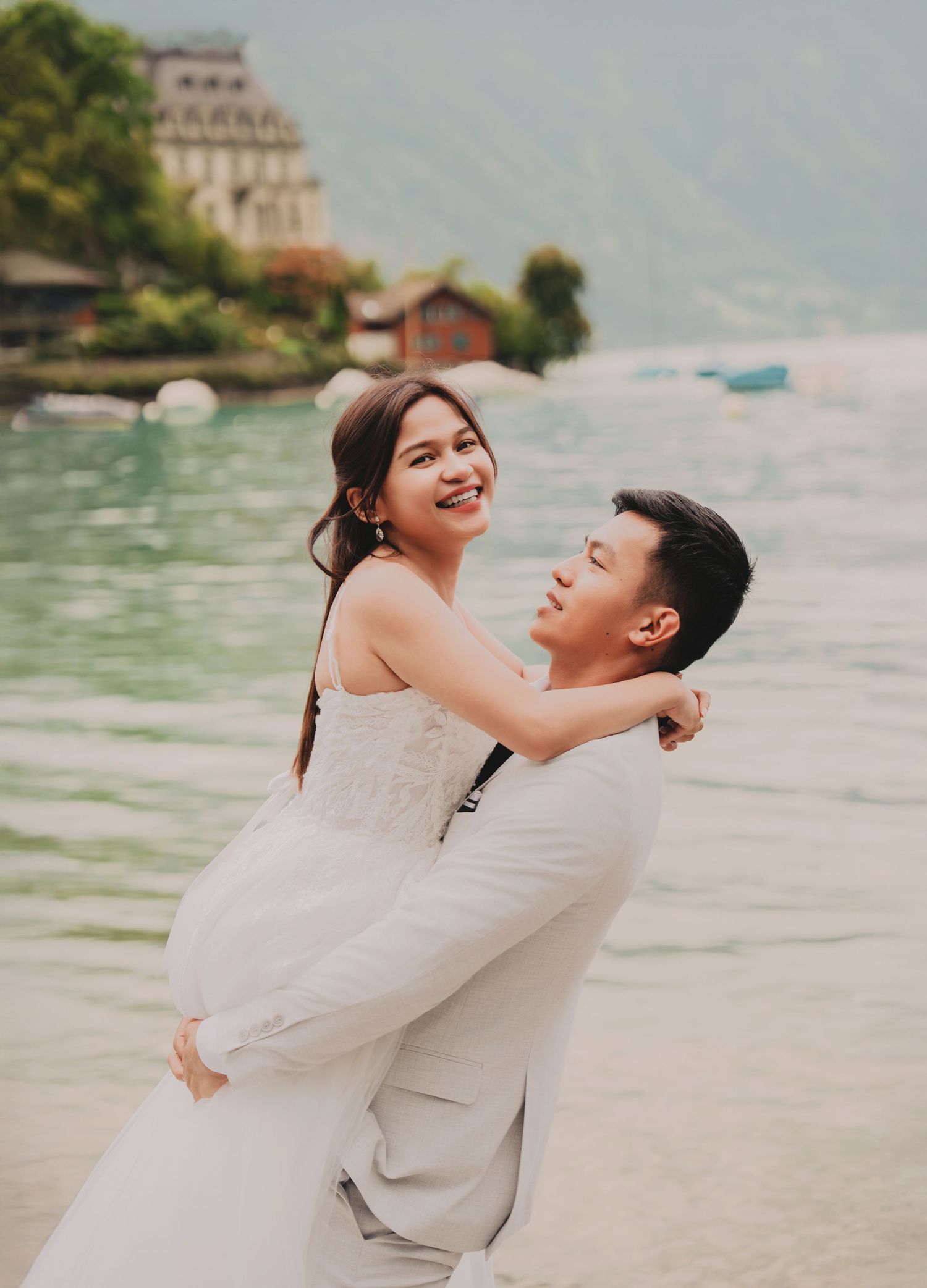 Groom lifting smiling bride by a stunning alpine lake with lush green mountains and historic building backdrop.