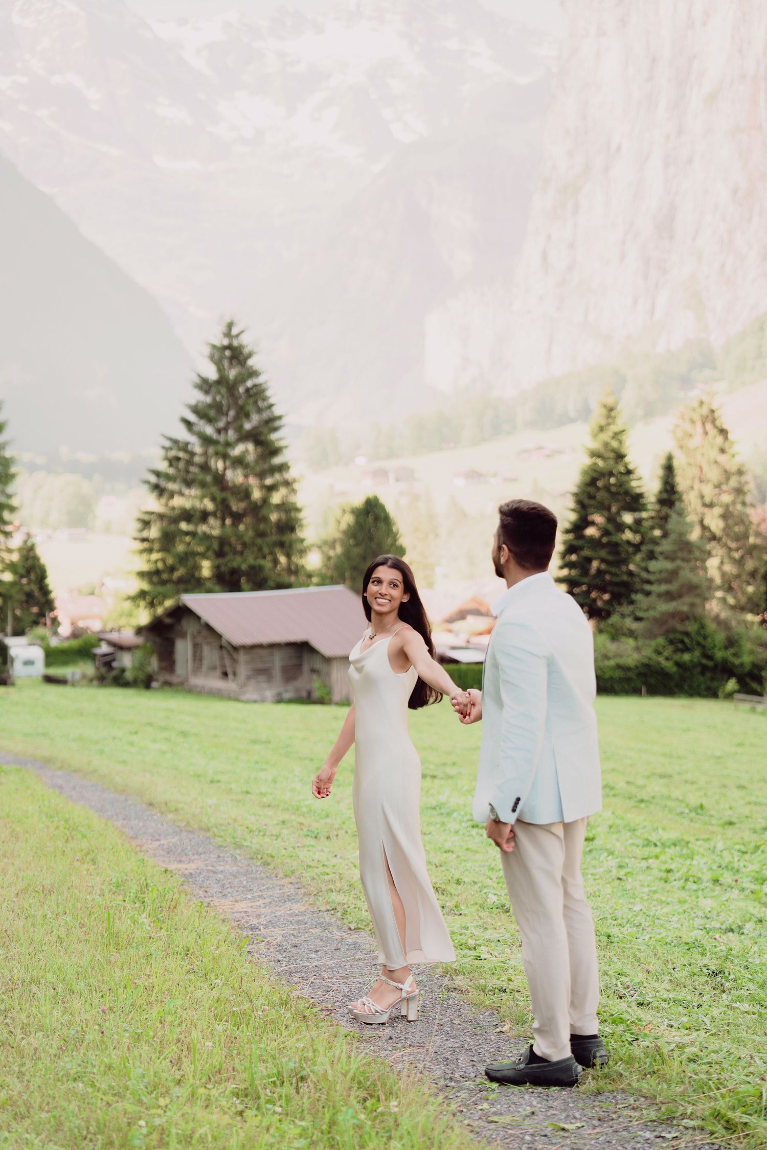 Joyful bride in white dress holding groom's hand while walking on path in Swiss valley.