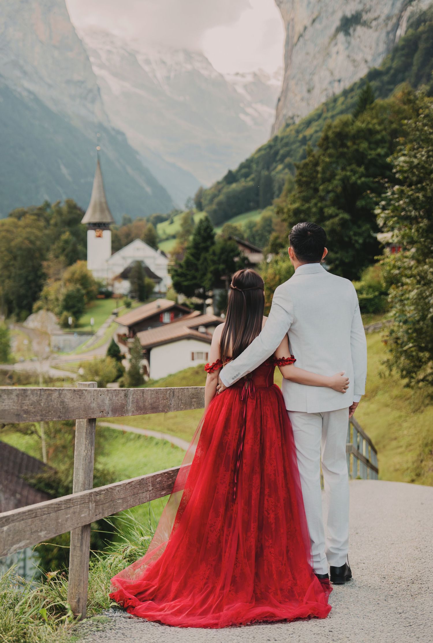 Couple from behind admiring Swiss mountain village view, woman in flowing red gown, man in white suit.
