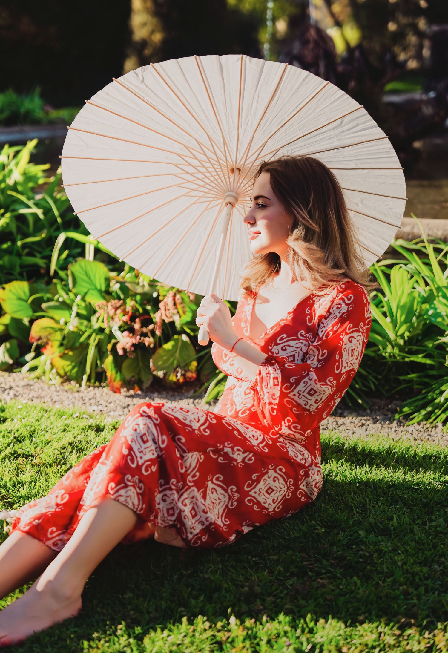 Woman in red patterned dress sits on grass holding a white parasol umbrella outdoors.