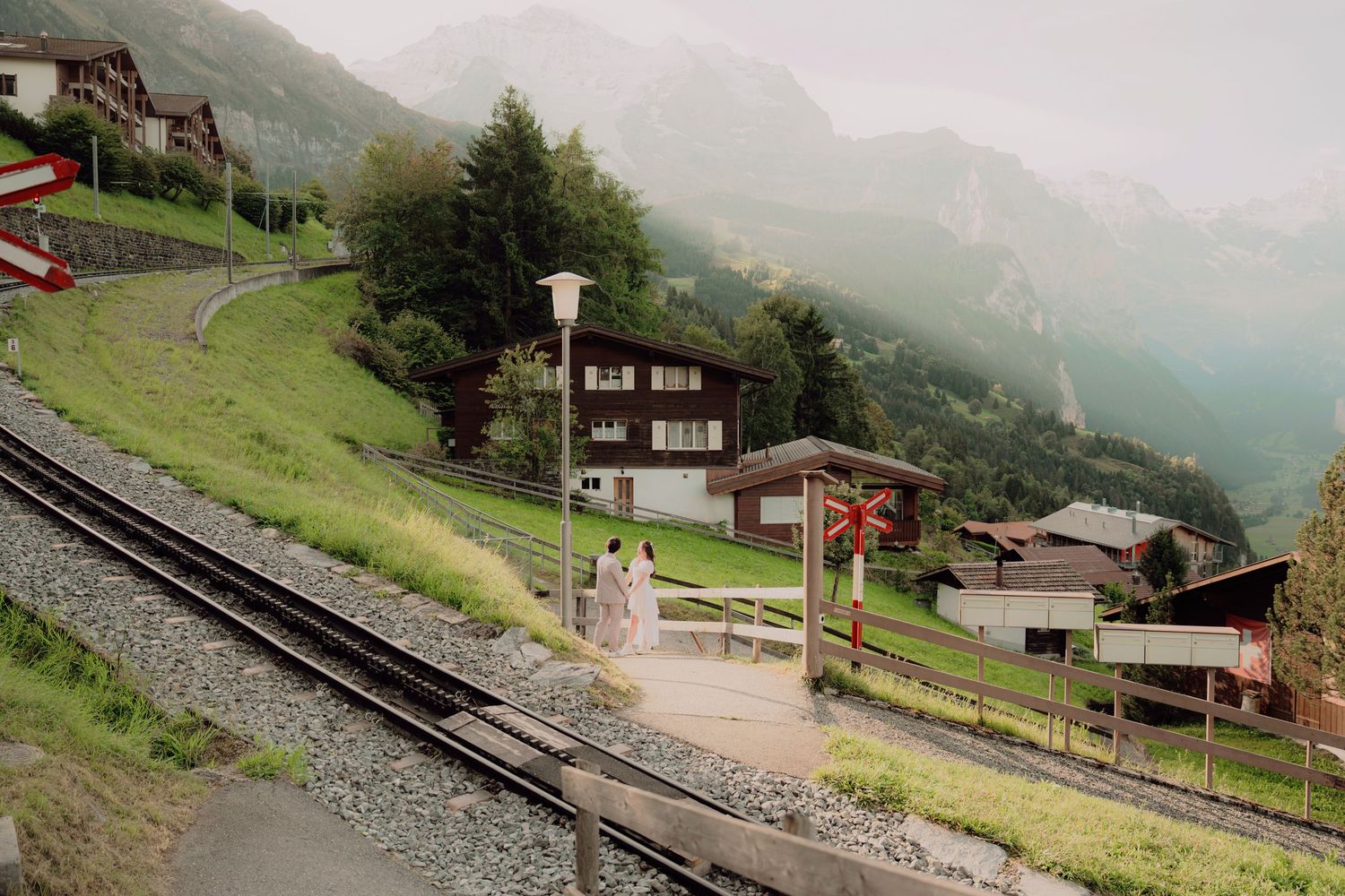 Wedding couple poses near a red railway crossing sign in a scenic Swiss alpine village with mountain backdrop.