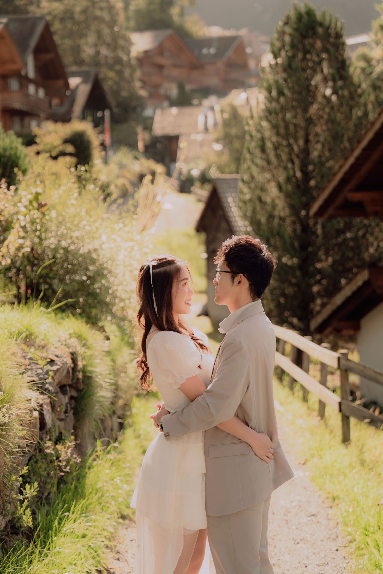 Couple embracing tenderly on sunlit path in Swiss alpine village with wooden chalets in soft background.