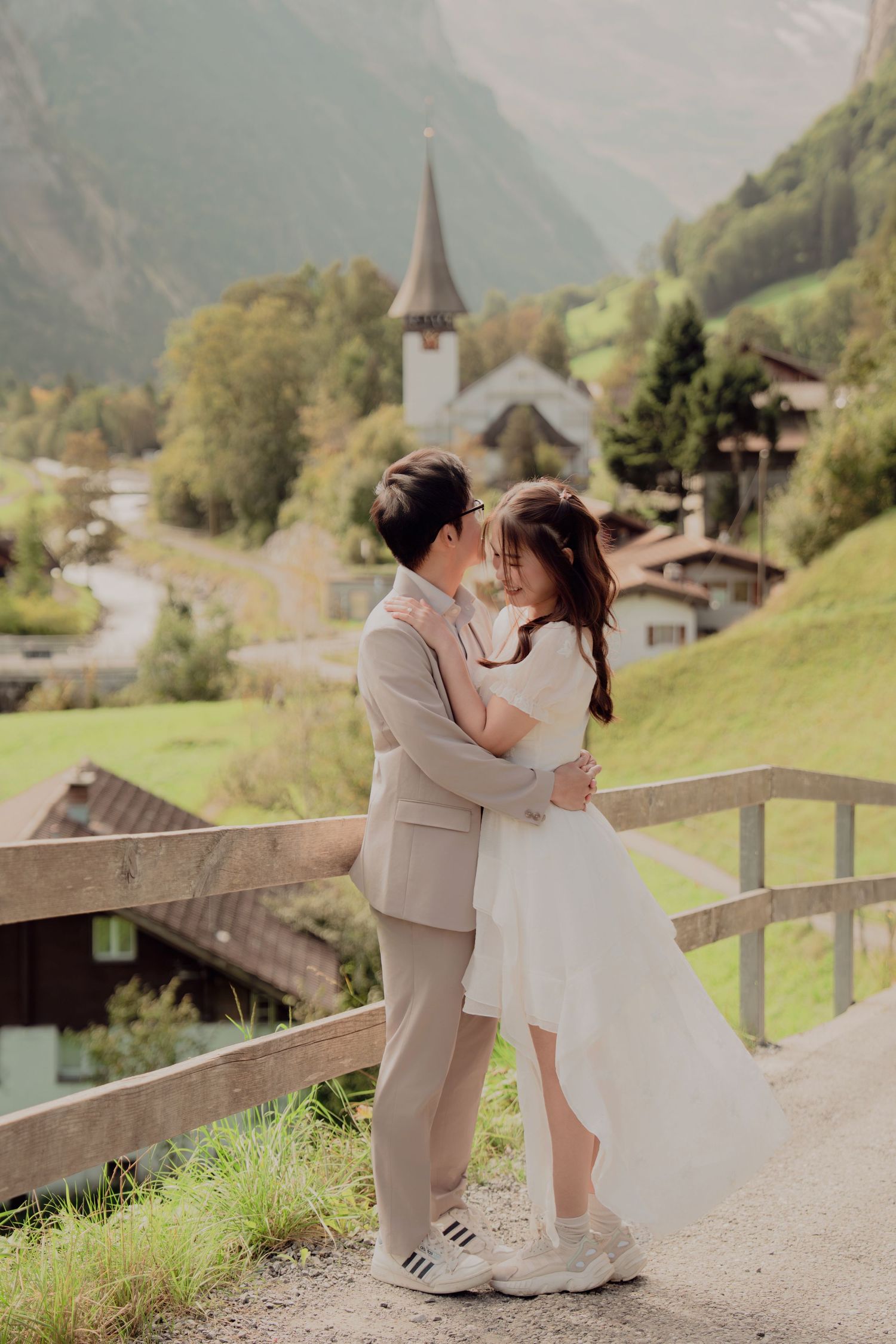 Couple sharing an intimate moment on a wooden fence path with Swiss alpine church village backdrop.