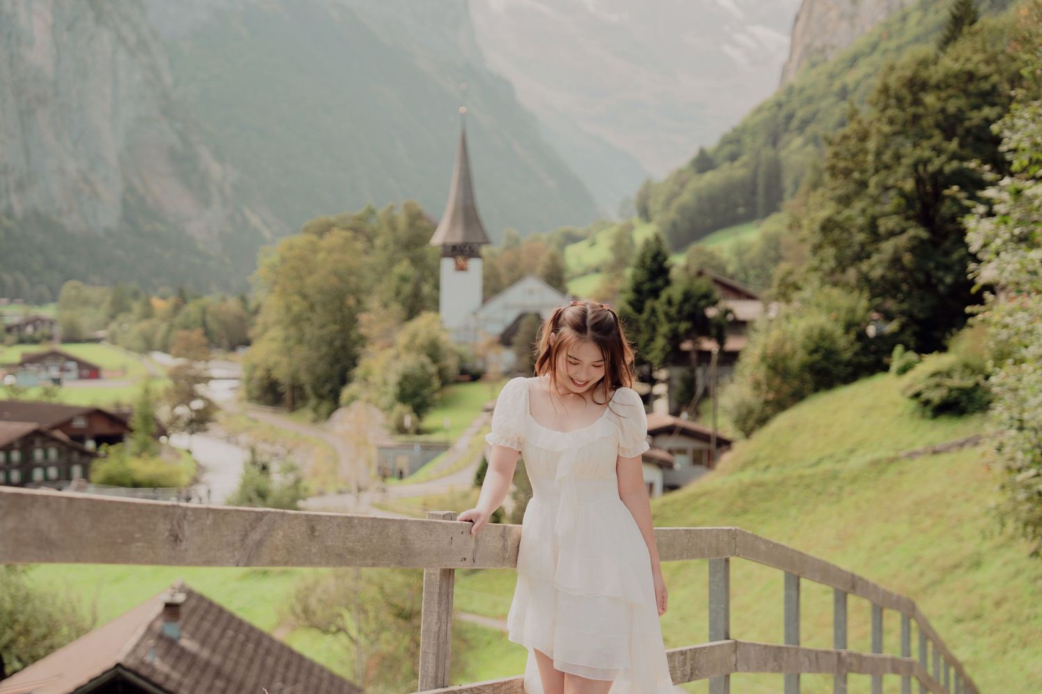Woman in white dress holding wooden railing on bridge with Swiss church and mountain village behind.