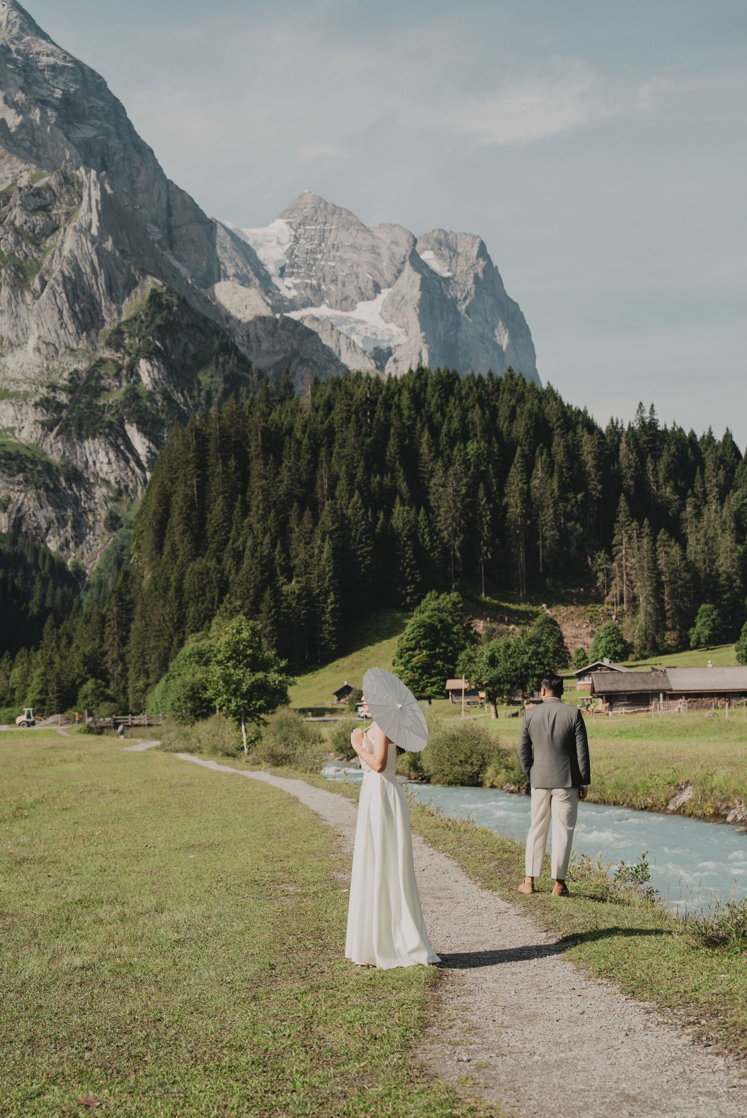 Elegant bride holding parasol and suited groom walk along mountain stream in scenic Swiss Alps.
