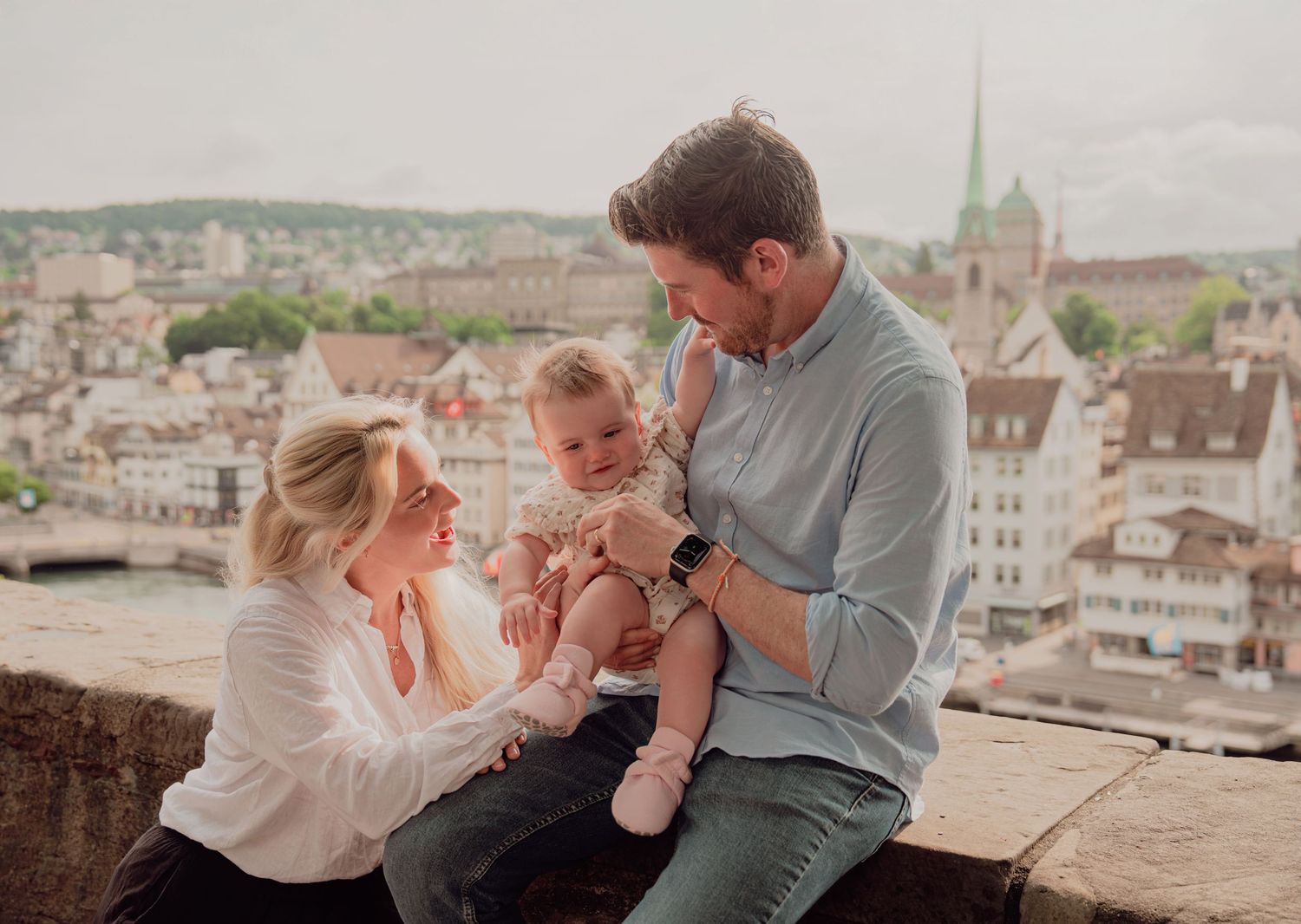 Happy family of three with baby sitting on stone wall overlooking Zurich cityscape.