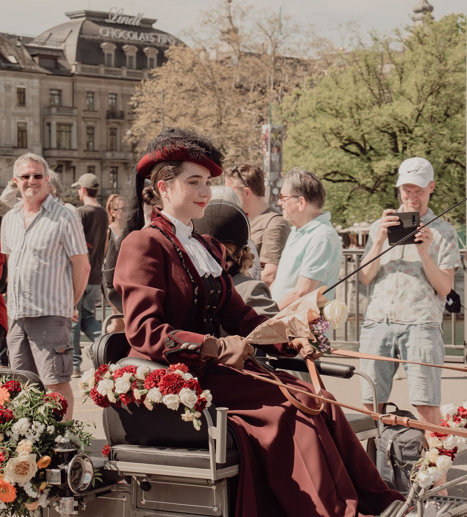 Elegant woman in burgundy Victorian dress driving a decorated carriage at a sunny parade.