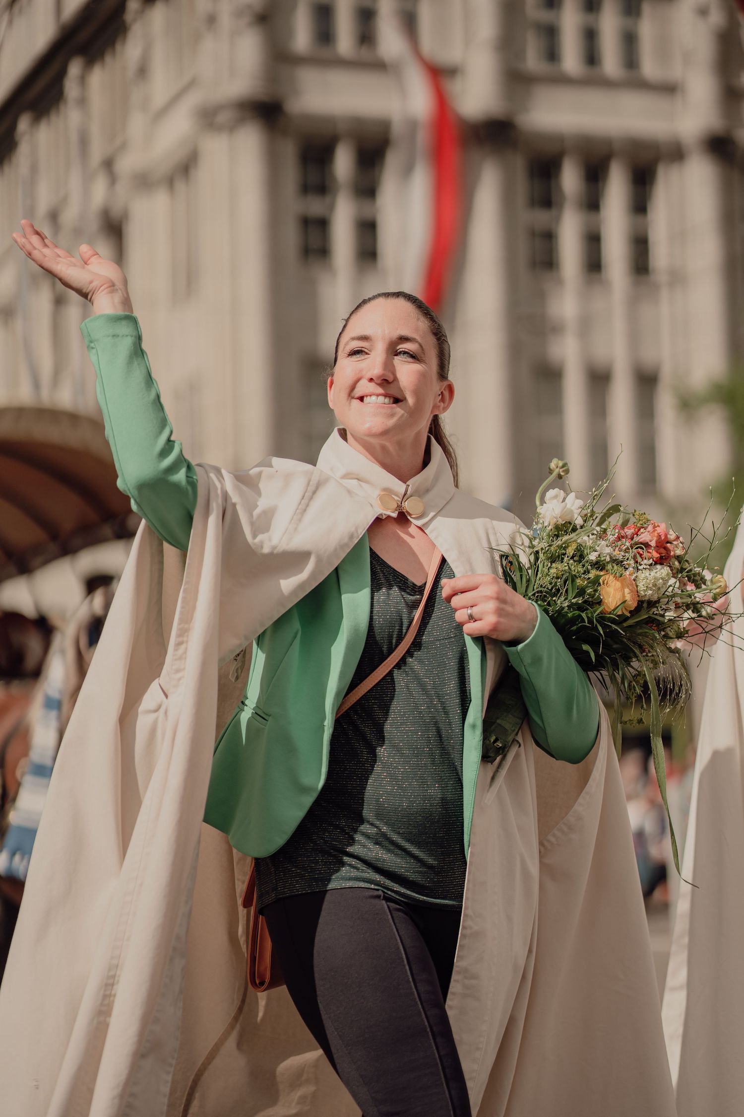 Smiling woman in green jacket and white cape waves while holding a bouquet at a street parade.