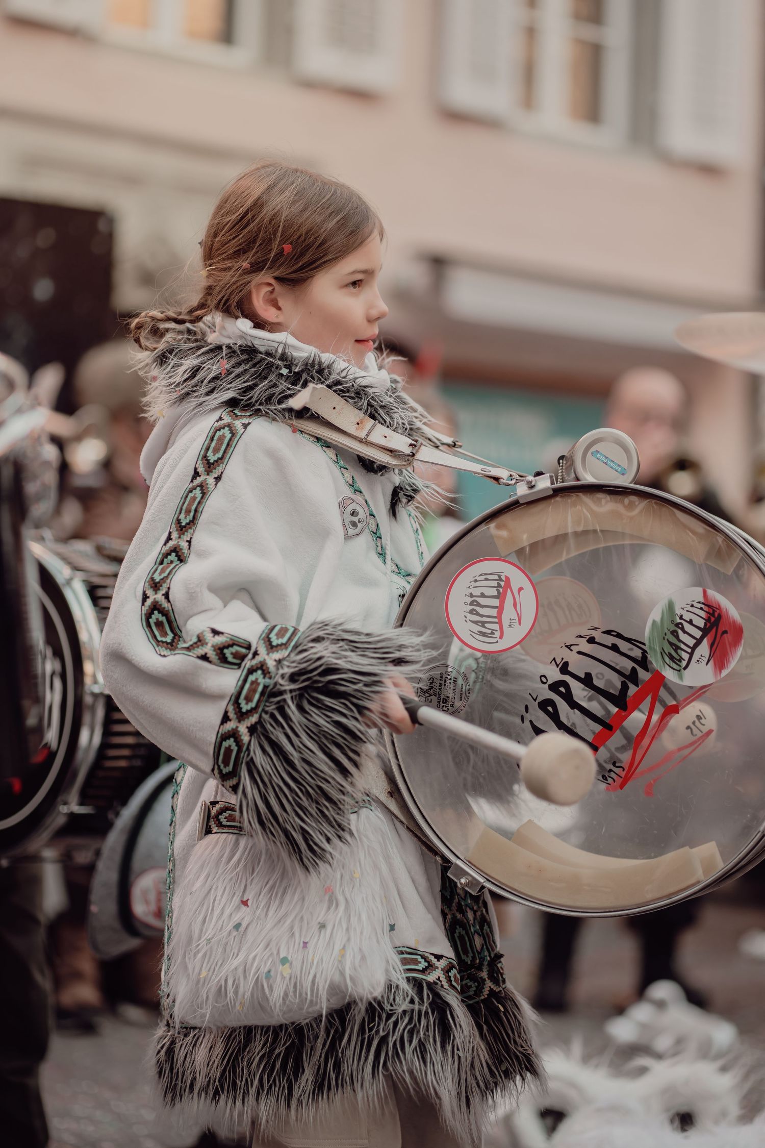 Young girl in decorative white fringed traditional costume playing a bass drum during a street parade.