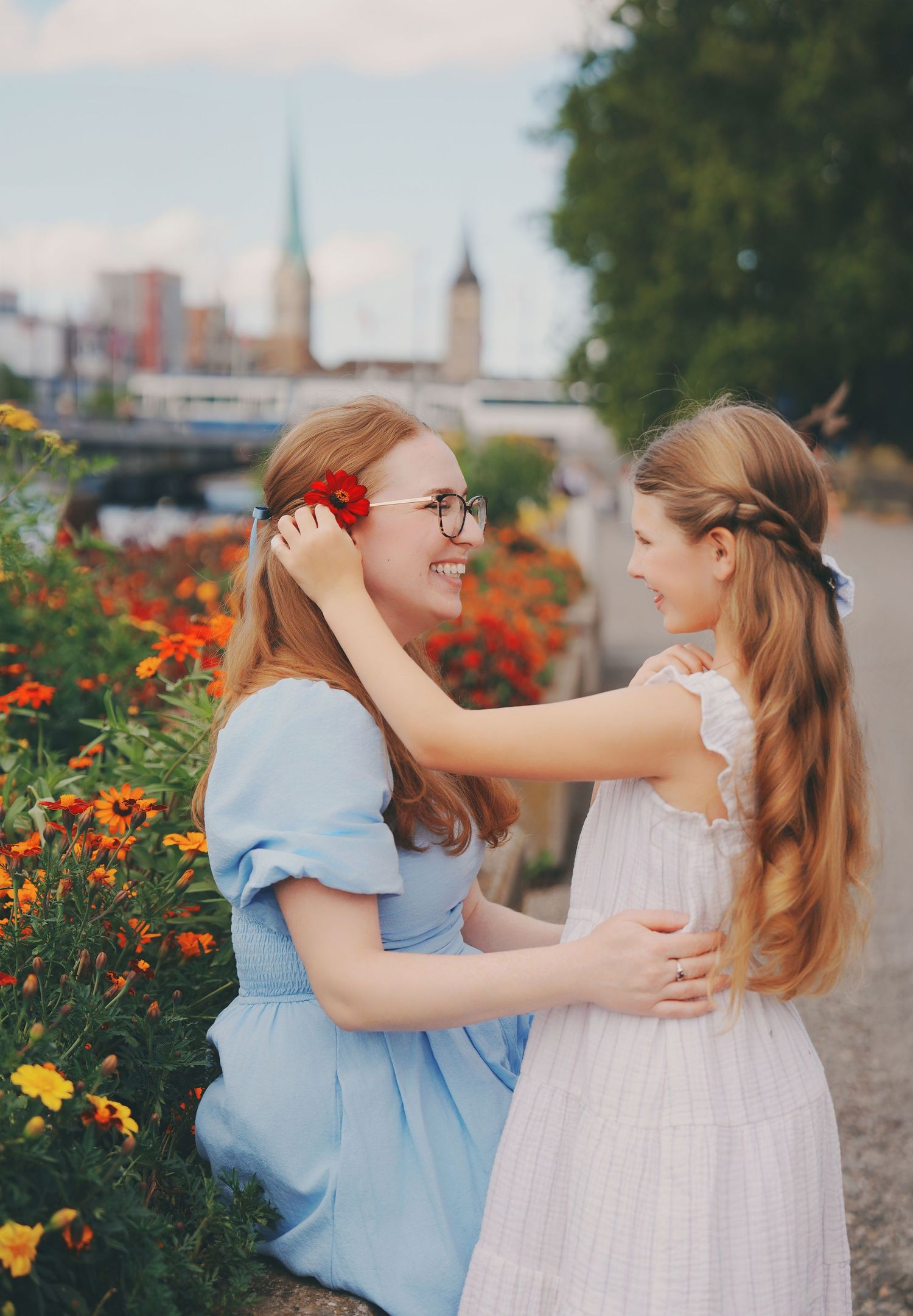Woman in blue dress smiling as girl places red flower in her hair by riverside.