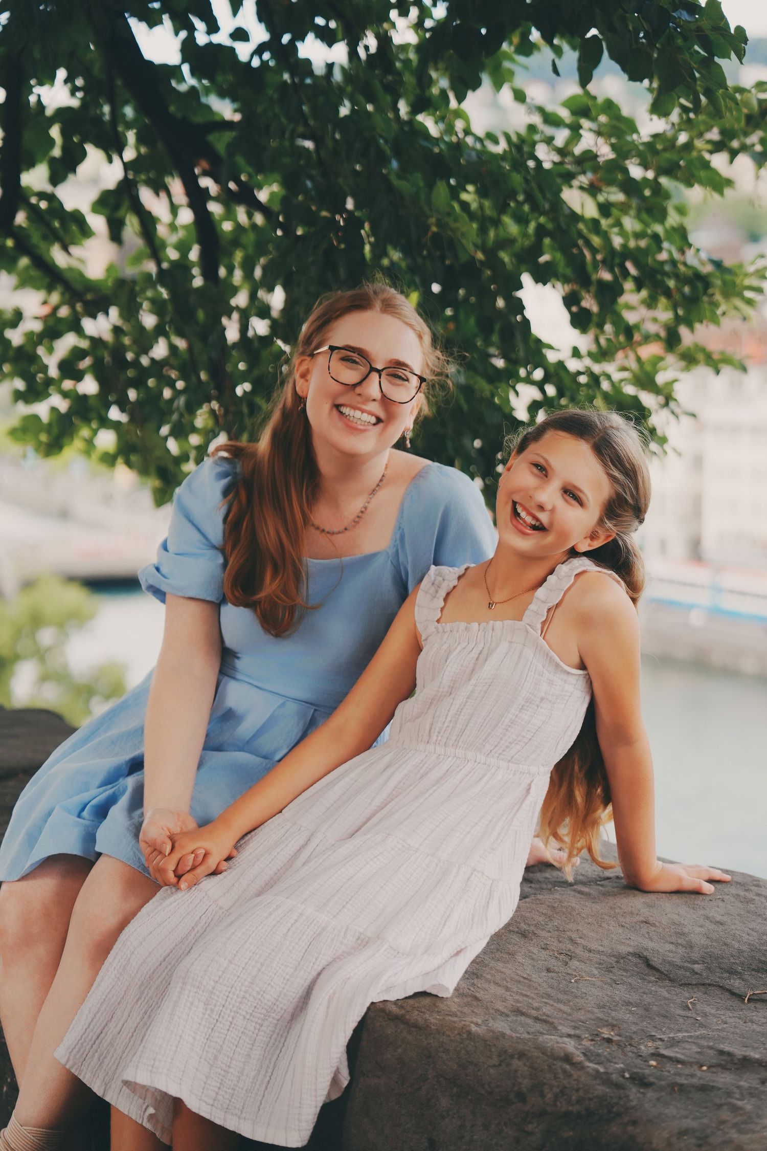 Two sisters laughing together seated on a stone wall surrounded by green trees.