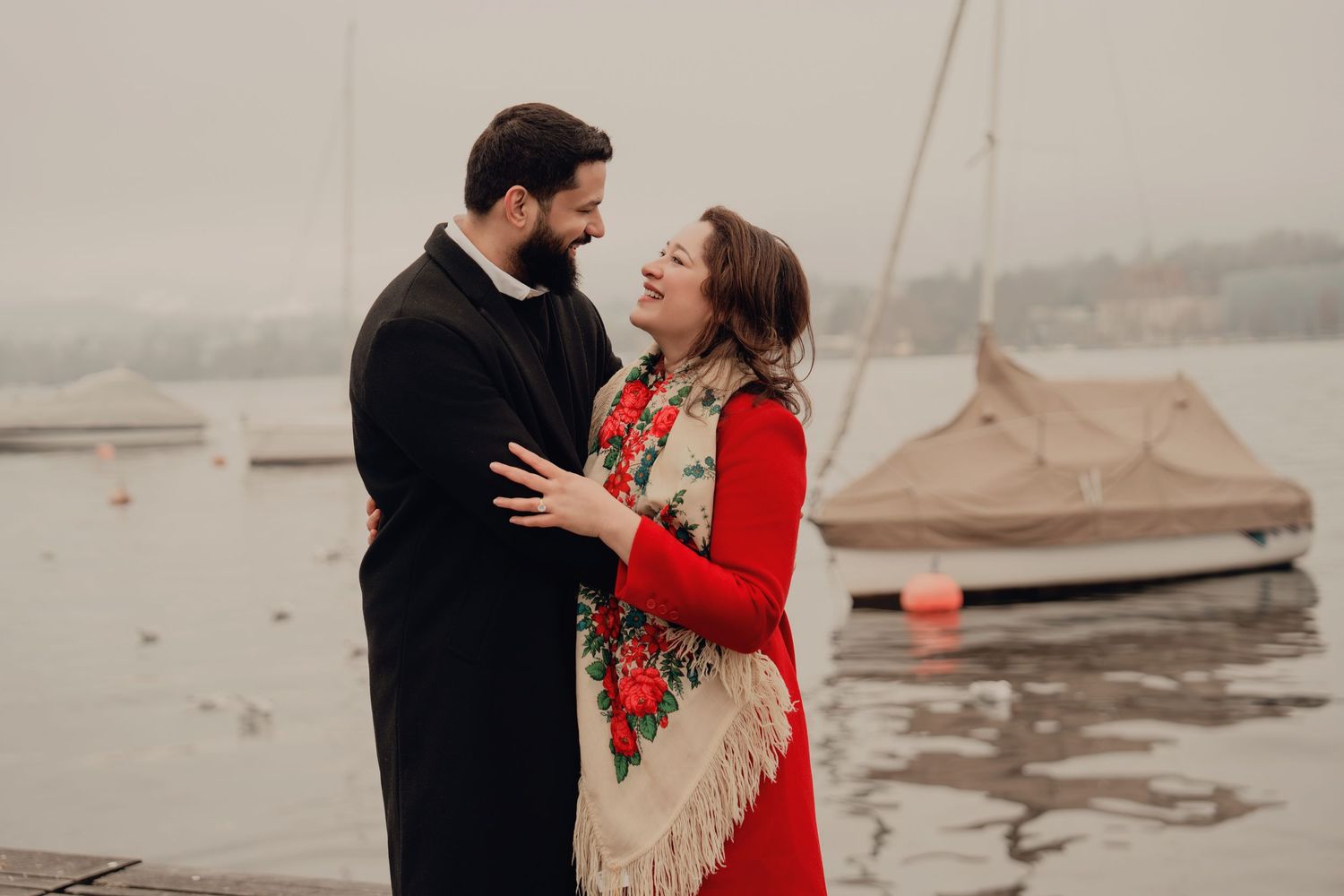 Romantic couple sharing a tender moment by the lake, woman in red coat and floral shawl, foggy backdrop.