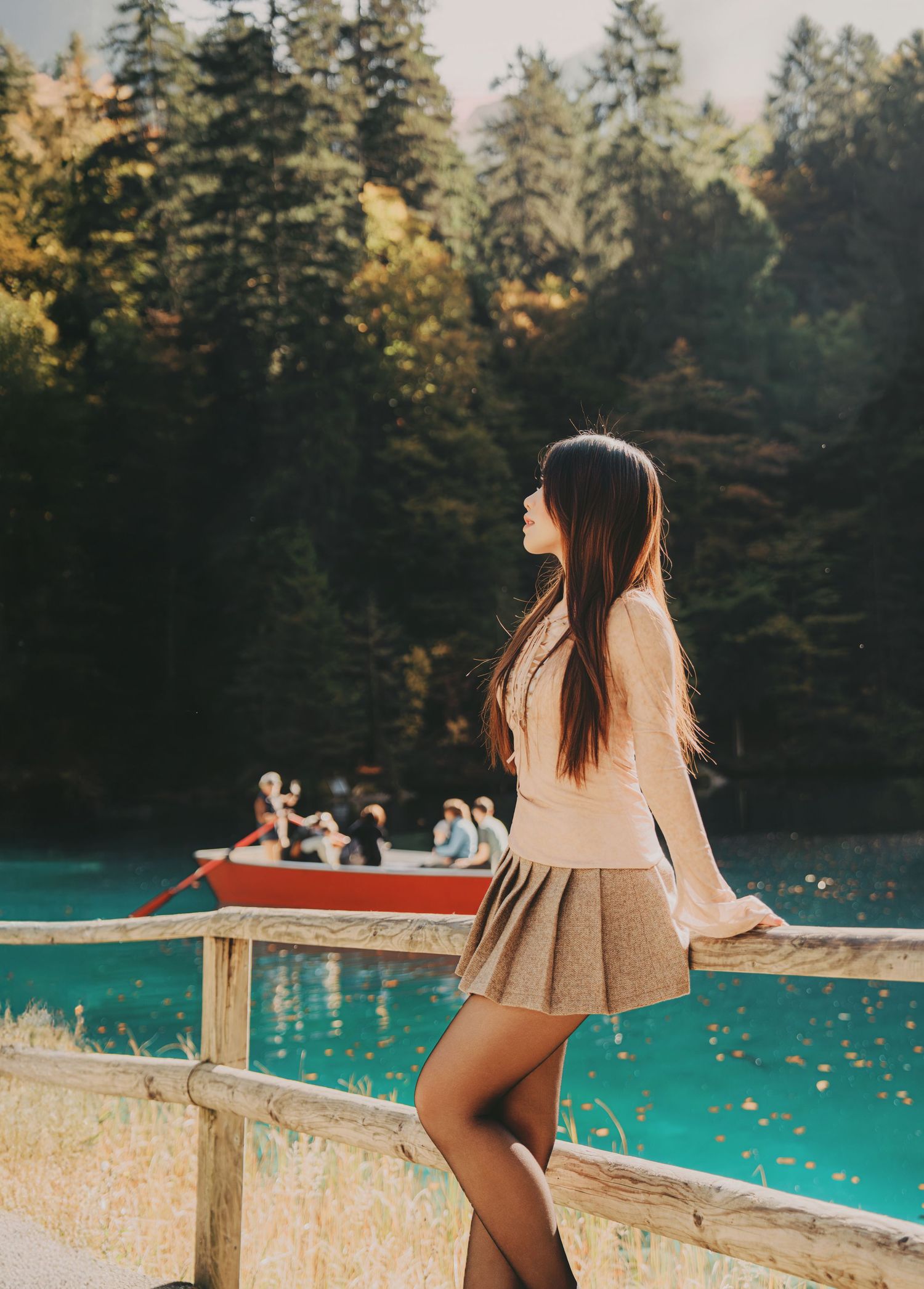 Young woman in beige top and gray skirt leans on wooden fence overlooking a turquoise lake.
